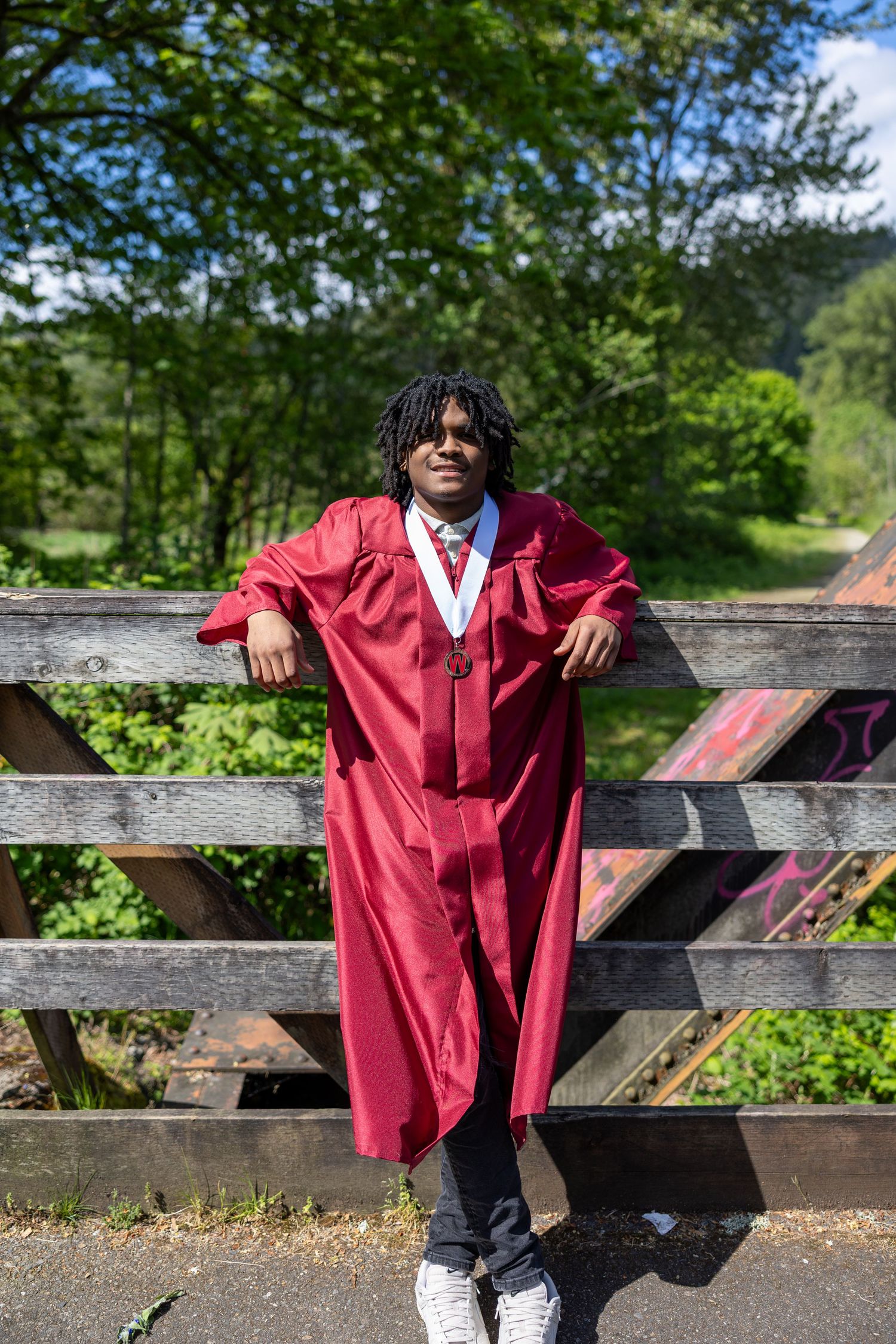 Graduate in red cap and gown leaning on wooden bridge railing surrounded by lush green trees.