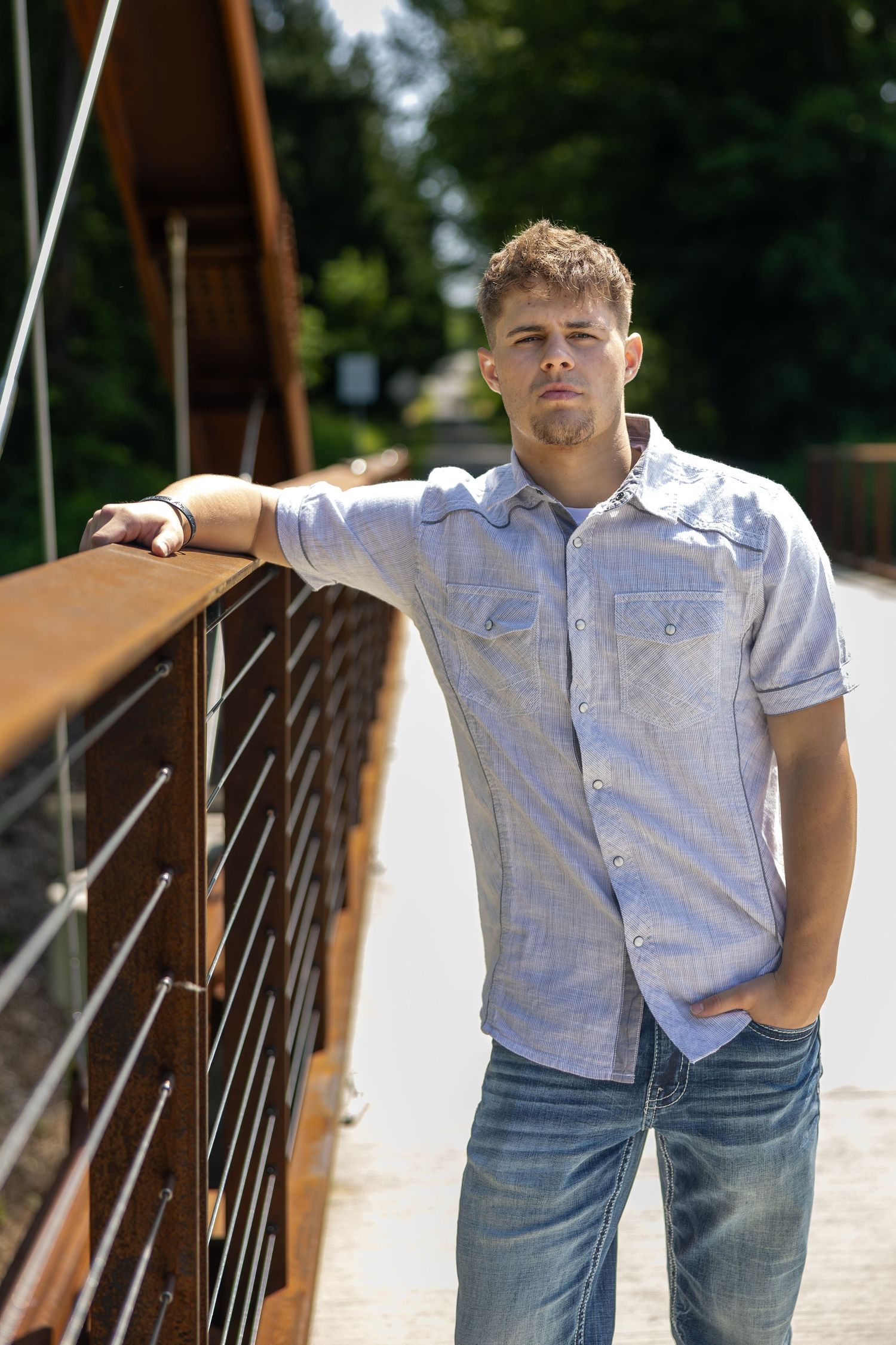 Young man in blue button-up shirt leaning on a bridge railing on a sunny day.