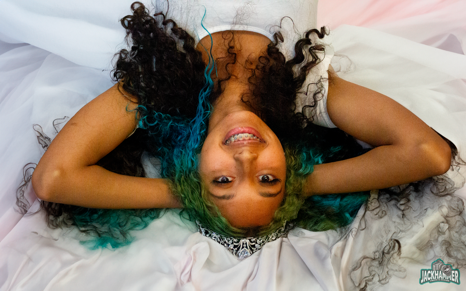 Top-down portrait of young woman with teal and black curly hair lying on white fabric, smiling.