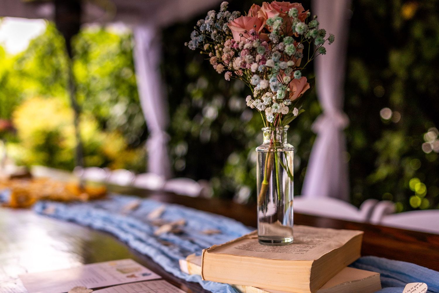 Glass bottle with colorful dried flowers placed on stacked books at an outdoor venue.