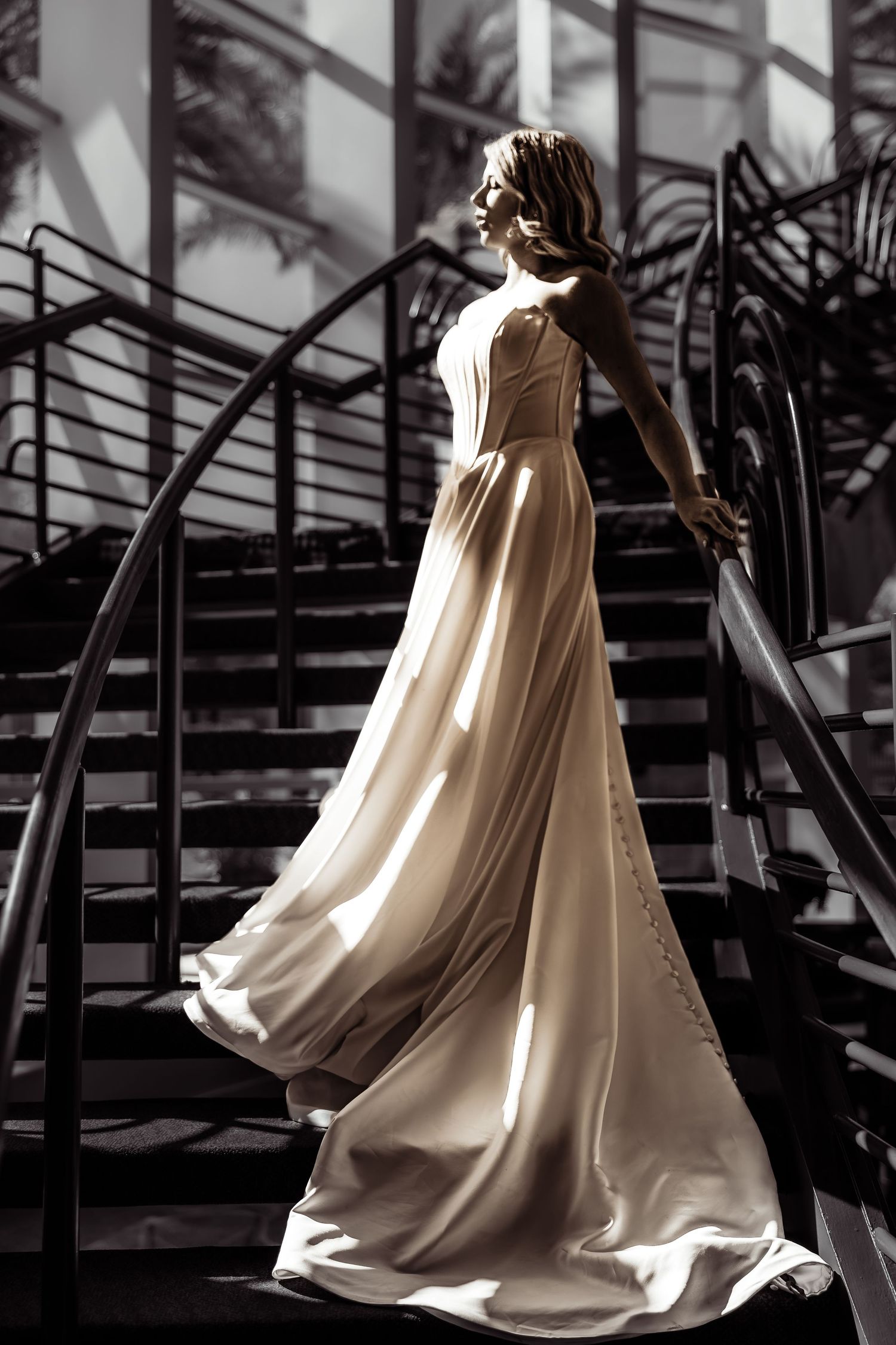Elegant bride in flowing white gown poses on ornate black staircase in dramatic lighting.