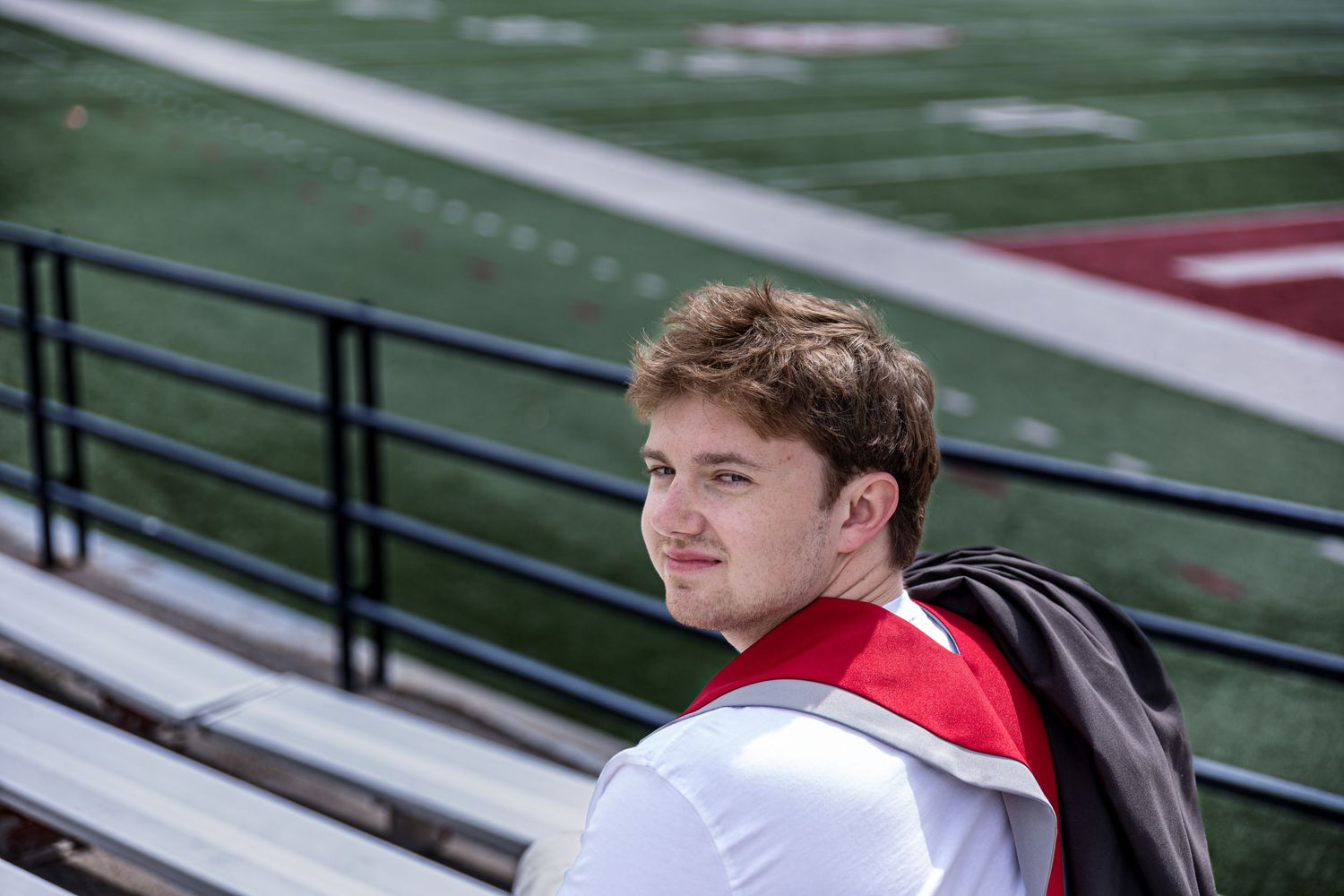 Teen boy in red and white uniform sitting in stadium bleachers looking over his shoulder.