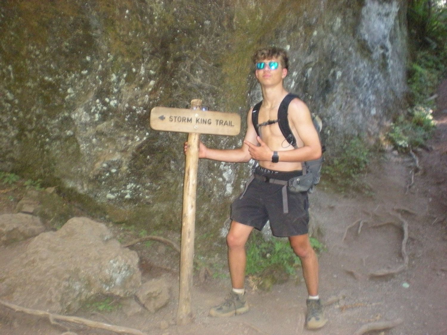 Hiker posing at Storm King Trail sign with rocky cliff backdrop wearing sunglasses and backpack.