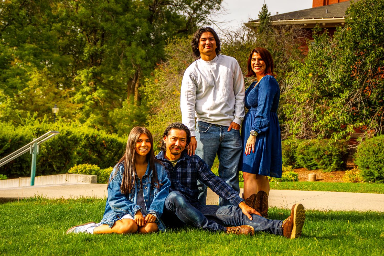 A smiling family of four poses together on green grass in a sunny park with trees in the background.