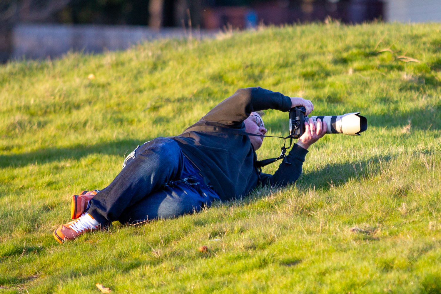 Photographer lying on grass capturing shots with a large telephoto lens camera in natural outdoor setting.