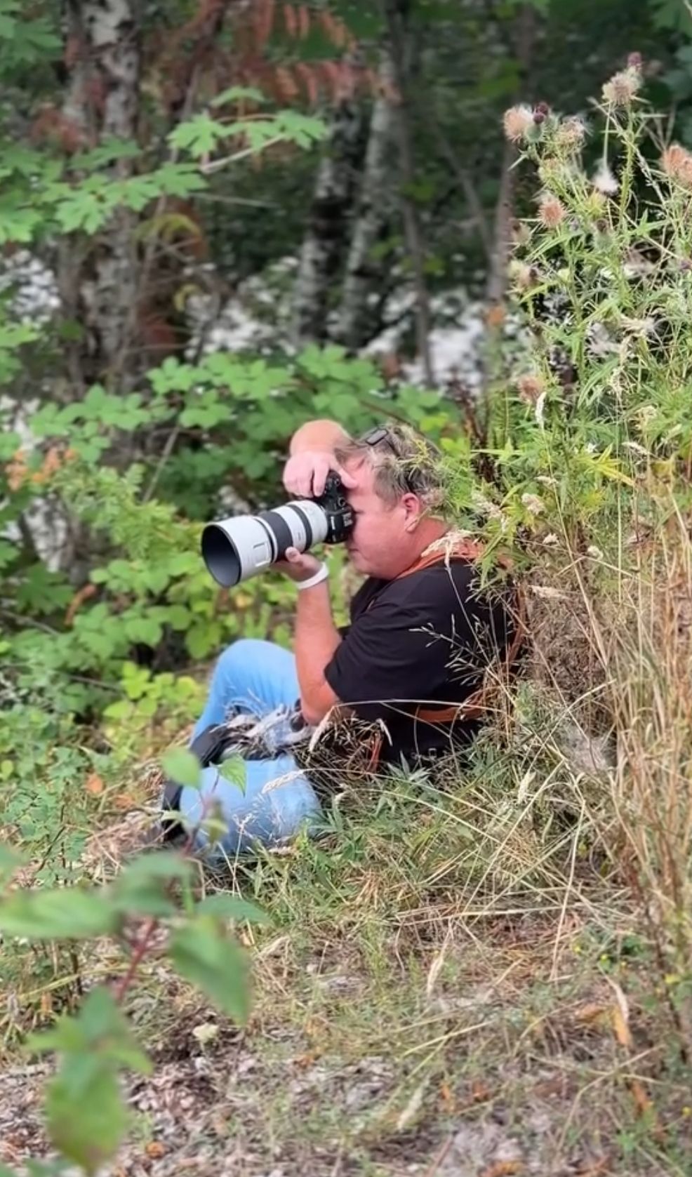 Photographer crouching in tall grass and bushes using a professional camera with a long telephoto lens.