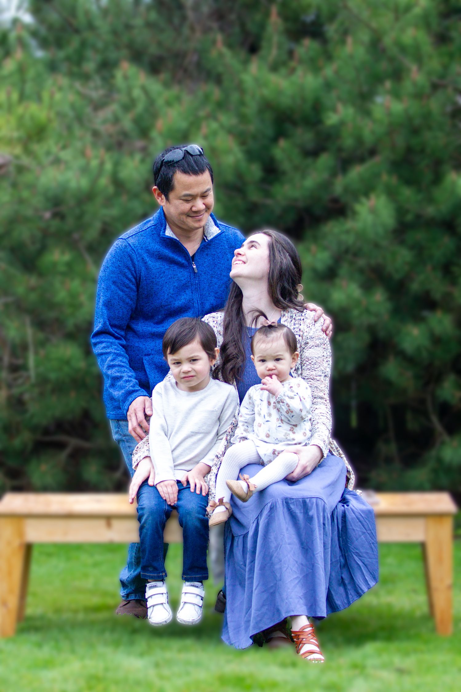 A happy family of four poses on a wooden bench surrounded by lush green trees in an outdoor setting.