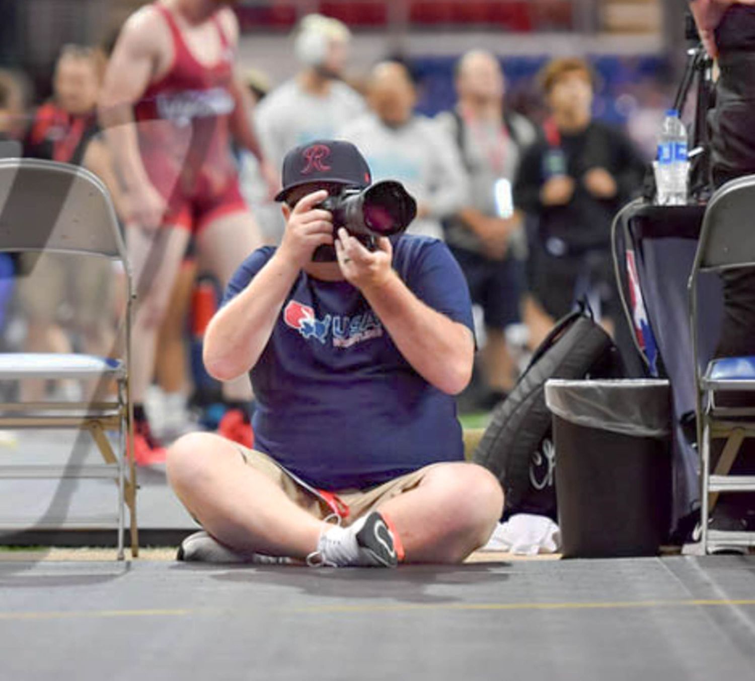 Sports photographer sitting cross-legged on the floor capturing action shots at an indoor wrestling event.