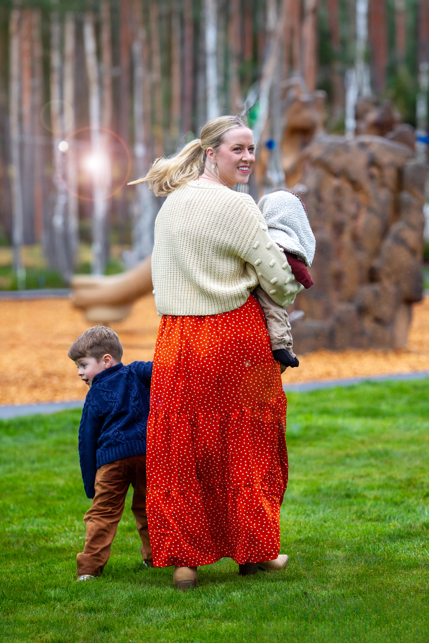 Blonde mother in red floral skirt carries baby while young boy walks beside her in park.