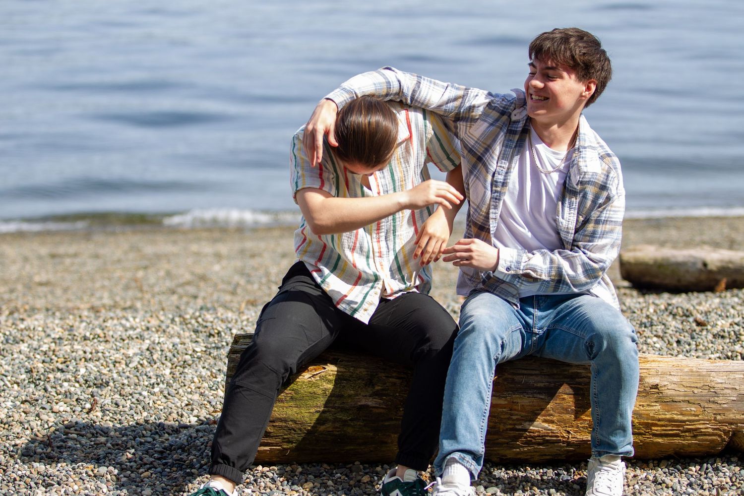 Two teenage boys sit on a driftwood log by a rocky beach, laughing and joking together.