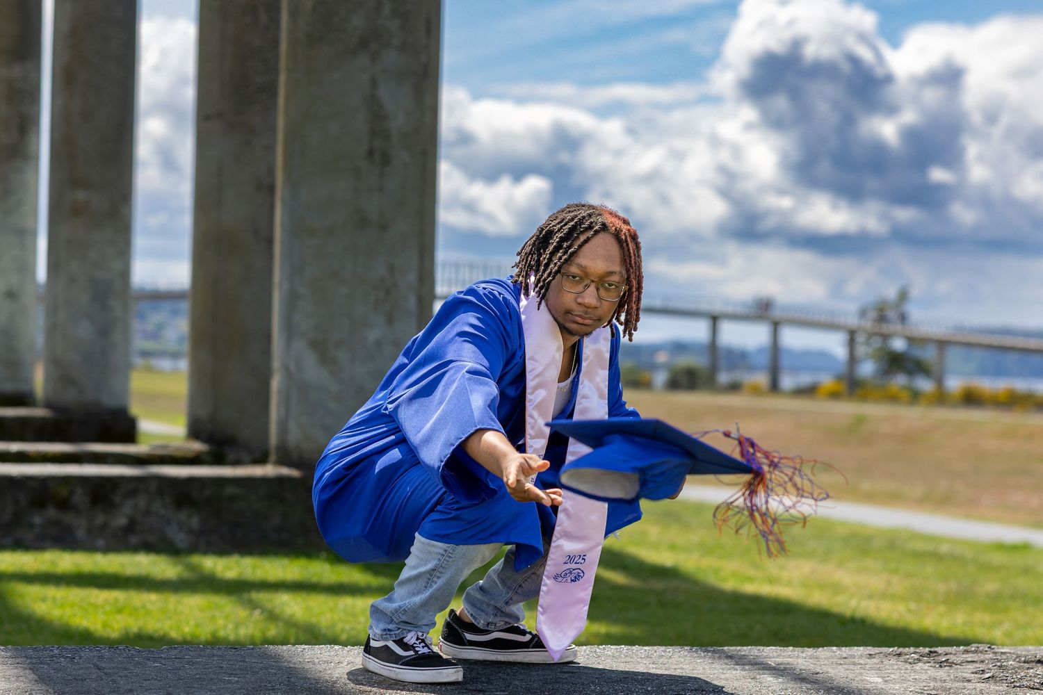 Young woman in blue graduation gown crouching outdoors near bridge columns on a sunny day.