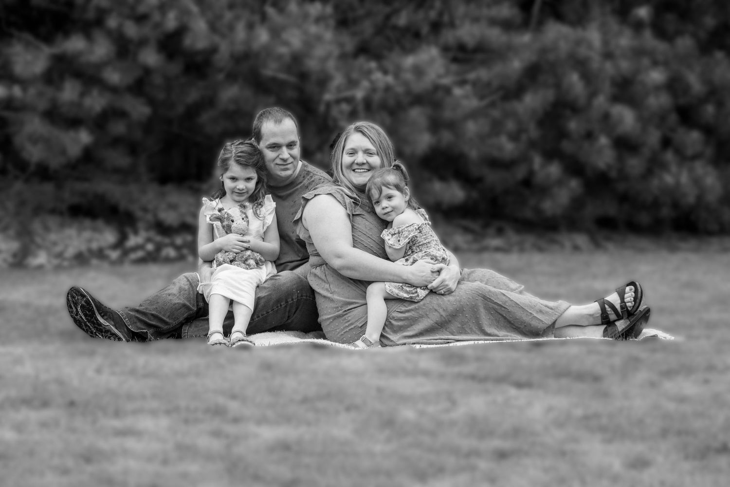 A black and white photo of a happy family of four sitting together on a blanket on a grassy lawn.