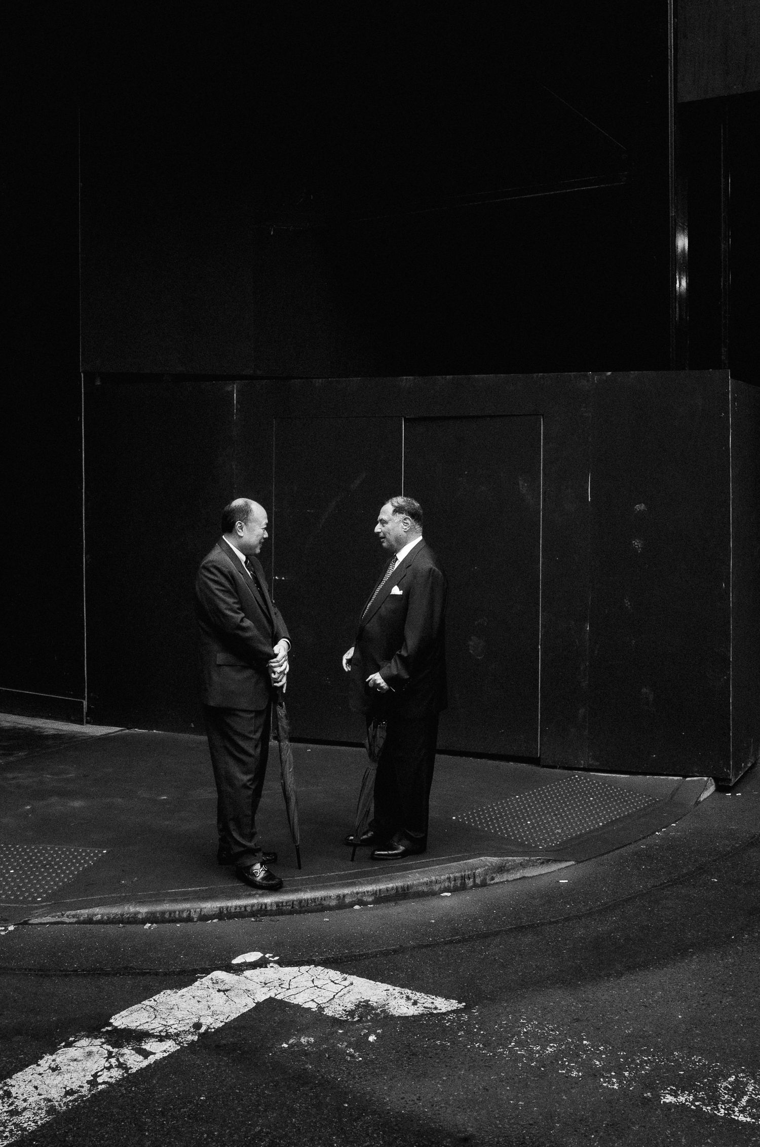 Two businessmen in suits having a conversation on a dark street corner with an arrow painted on the pavement.
