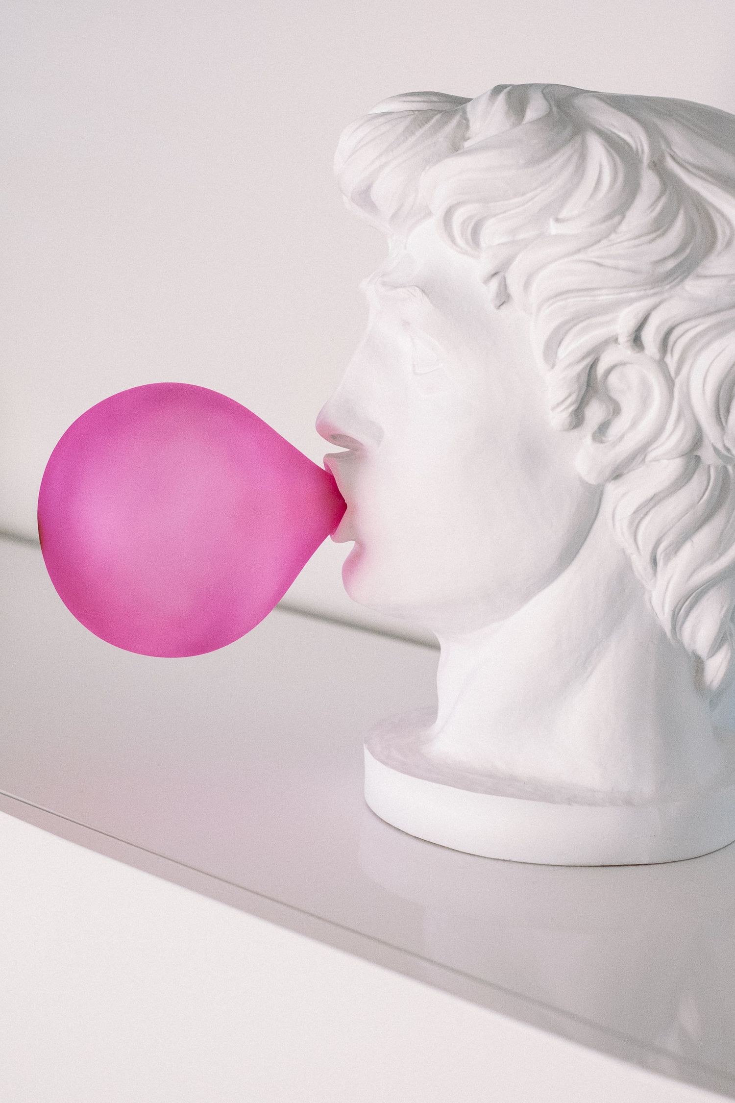 White classical sculpture bust with pink balloon creating artistic contrast against light background.