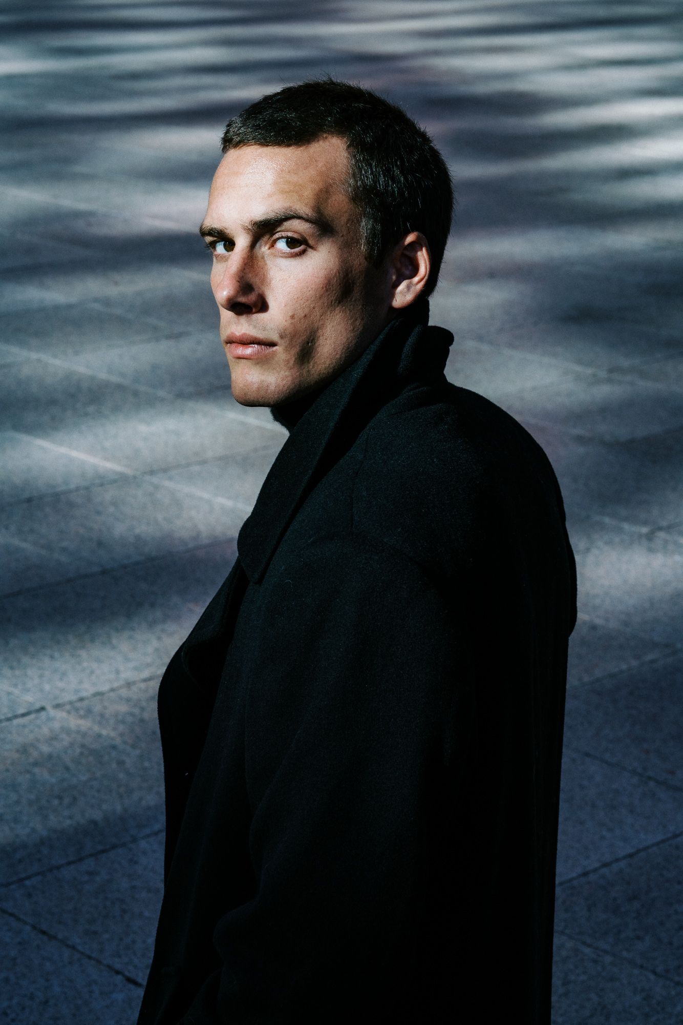 Dramatic portrait against blurred background wearing black coat in moody lighting.