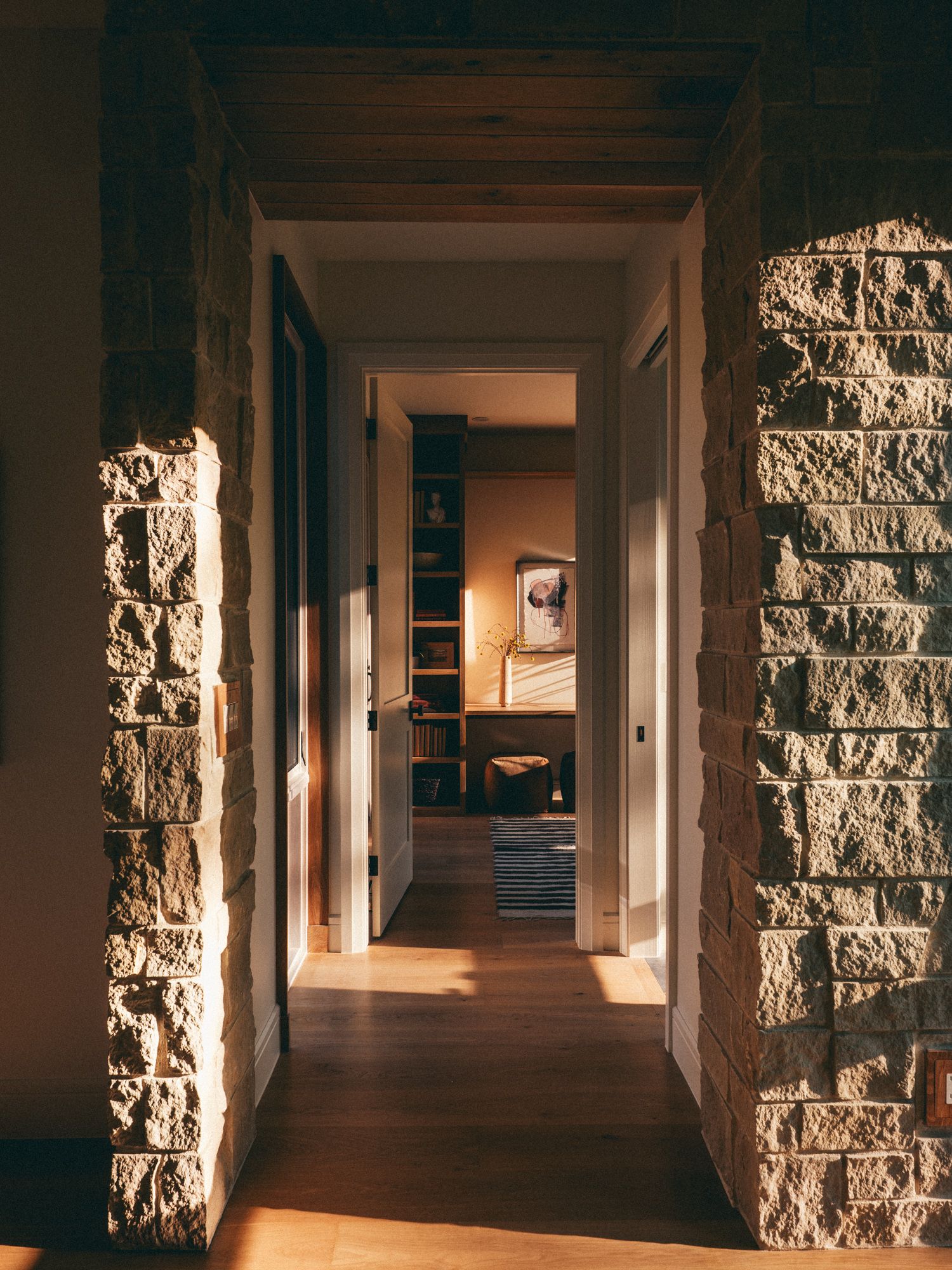 Sunlit hallway with rustic stone walls and hardwood floors leading to a warmly lit living room.