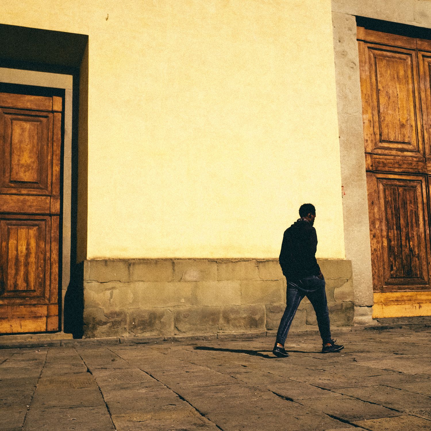 A silhouetted figure walks past wooden doors on a stone-paved street with beige walls in warm sunlight.