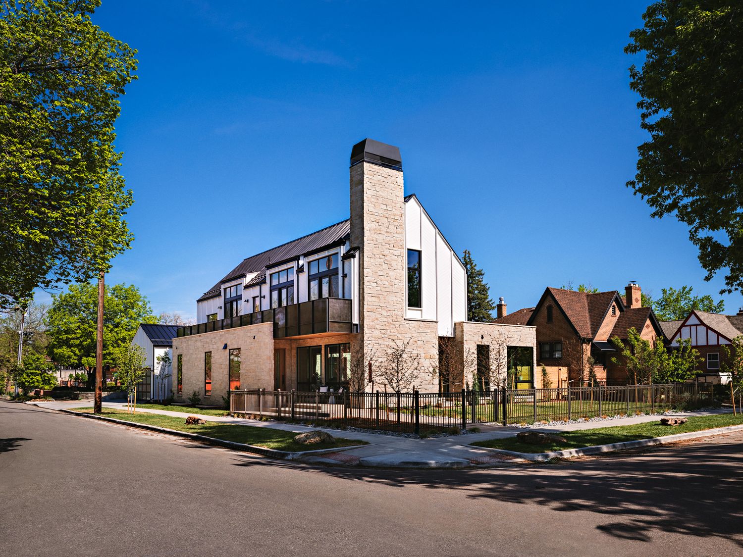 Modern brick and glass corner home with white chimney, black fence, and lush trees under a clear blue sky.