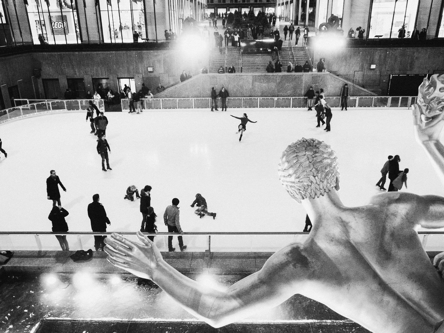 Black and white photograph of people ice skating on a rink with dramatic lighting.