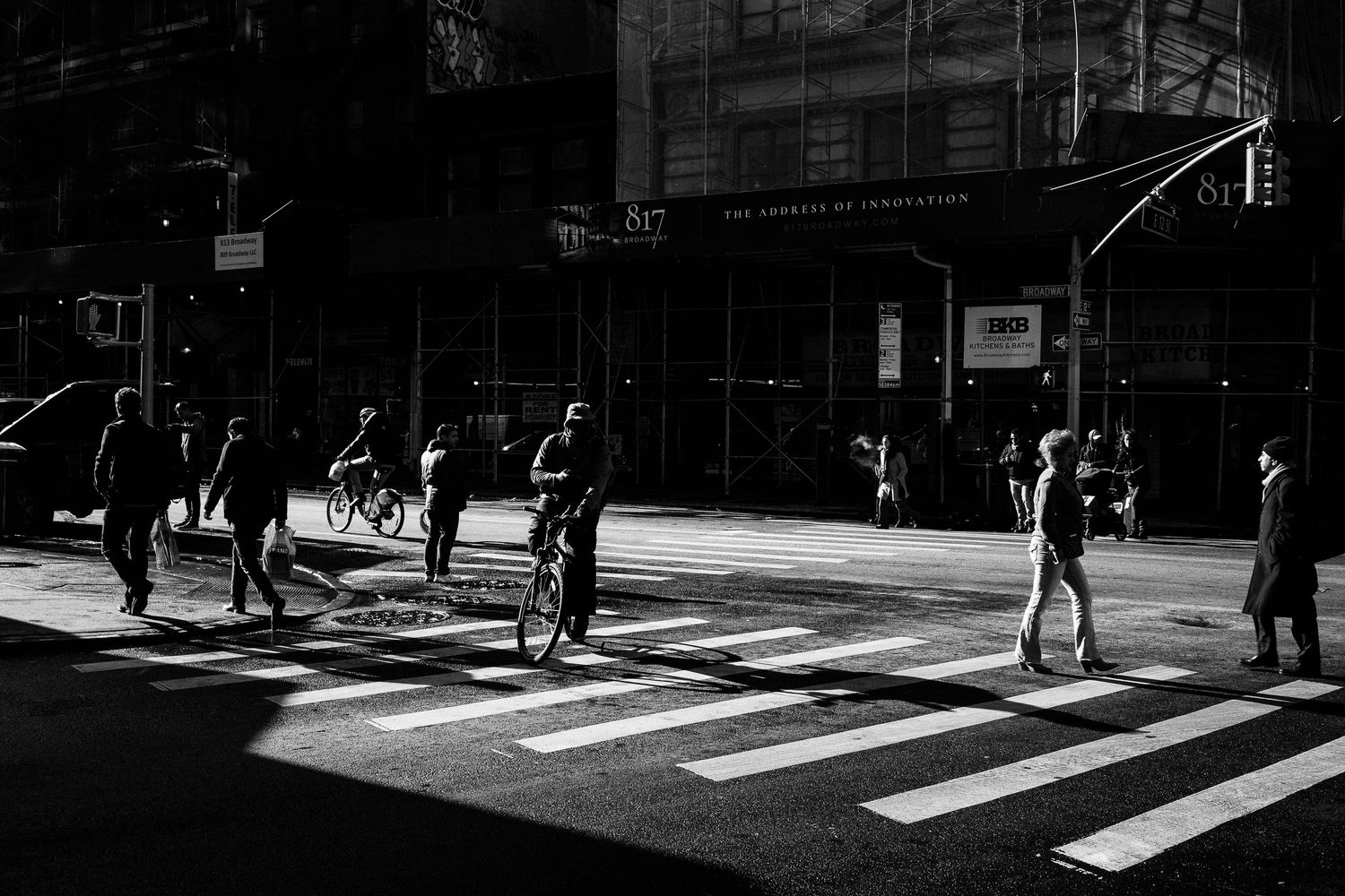Pedestrians and cyclists cross a sunlit intersection in a dramatic black and white urban street scene.