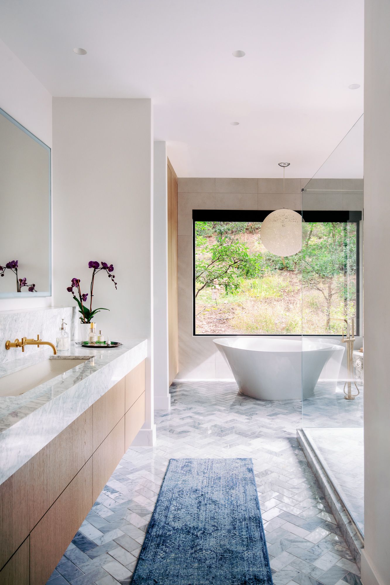 Elegant bathroom with marble herringbone floors, freestanding tub, and large window overlooking nature.