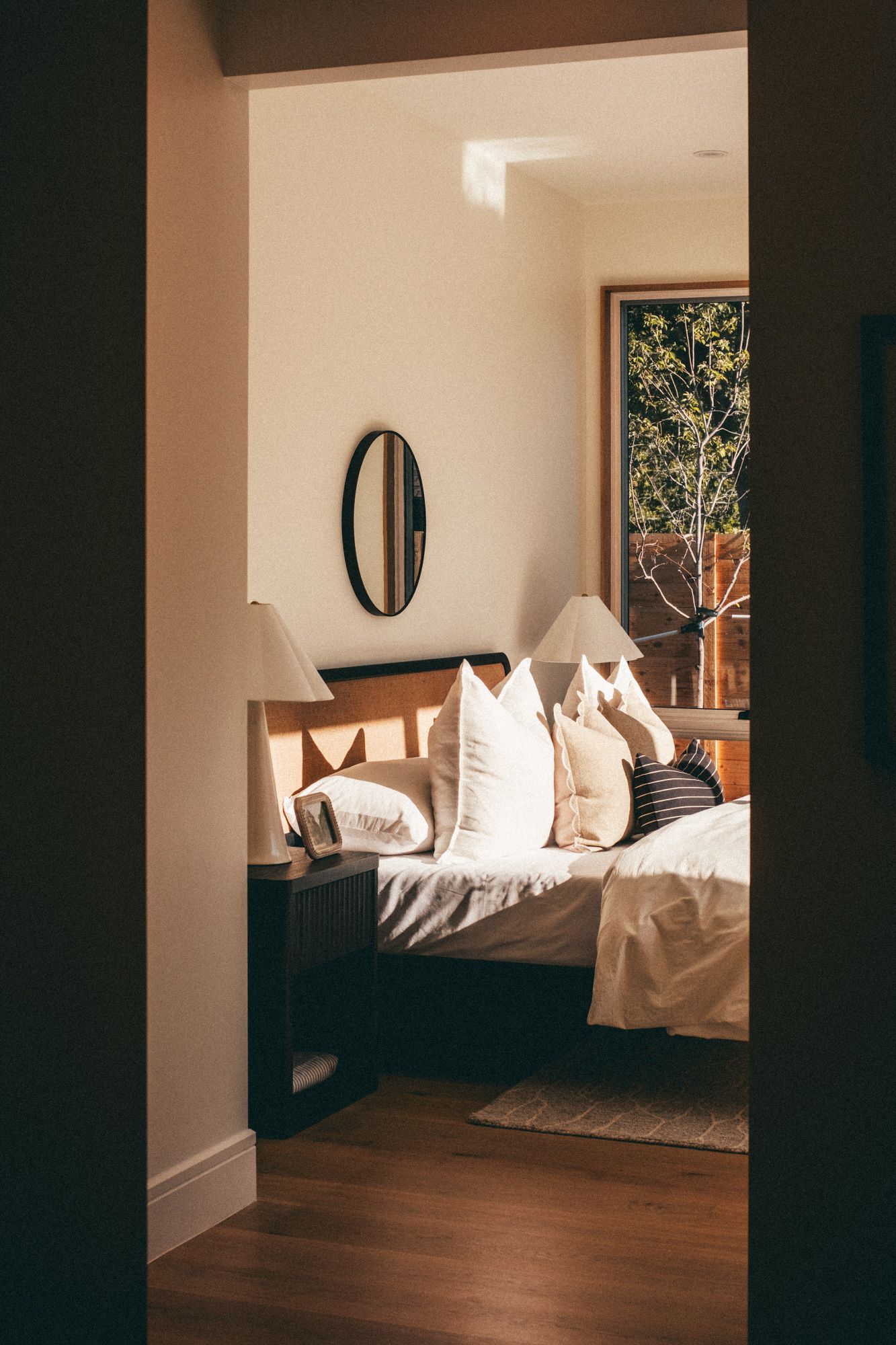 Elegant bedroom interior viewed through doorway with warm lighting, white pillows, oval mirror, and wood furniture.