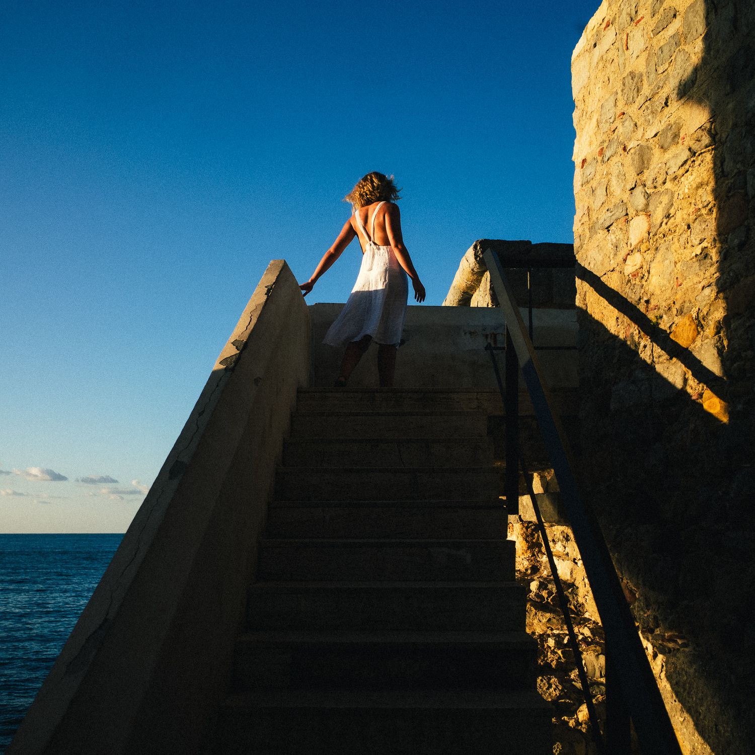 Silhouetted figures climb steep rocky cliffs against a dramatic blue sky and ocean backdrop at sunset.
