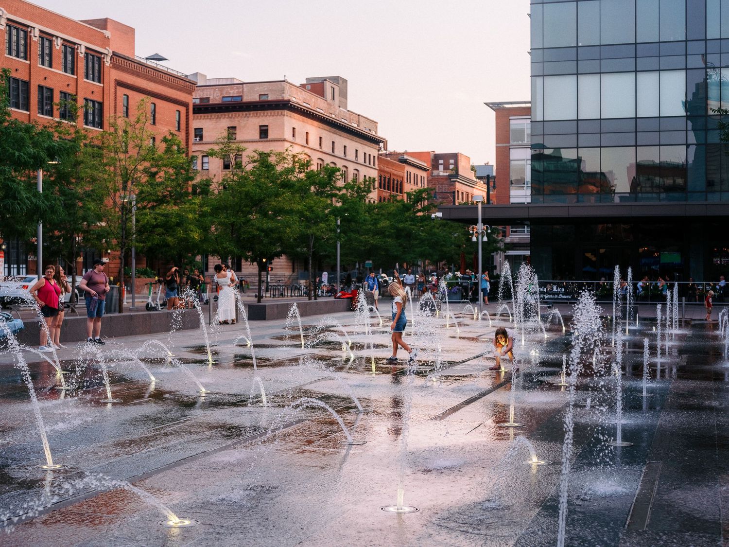 Interactive water fountains create a playful display in an urban plaza surrounded by brick buildings.