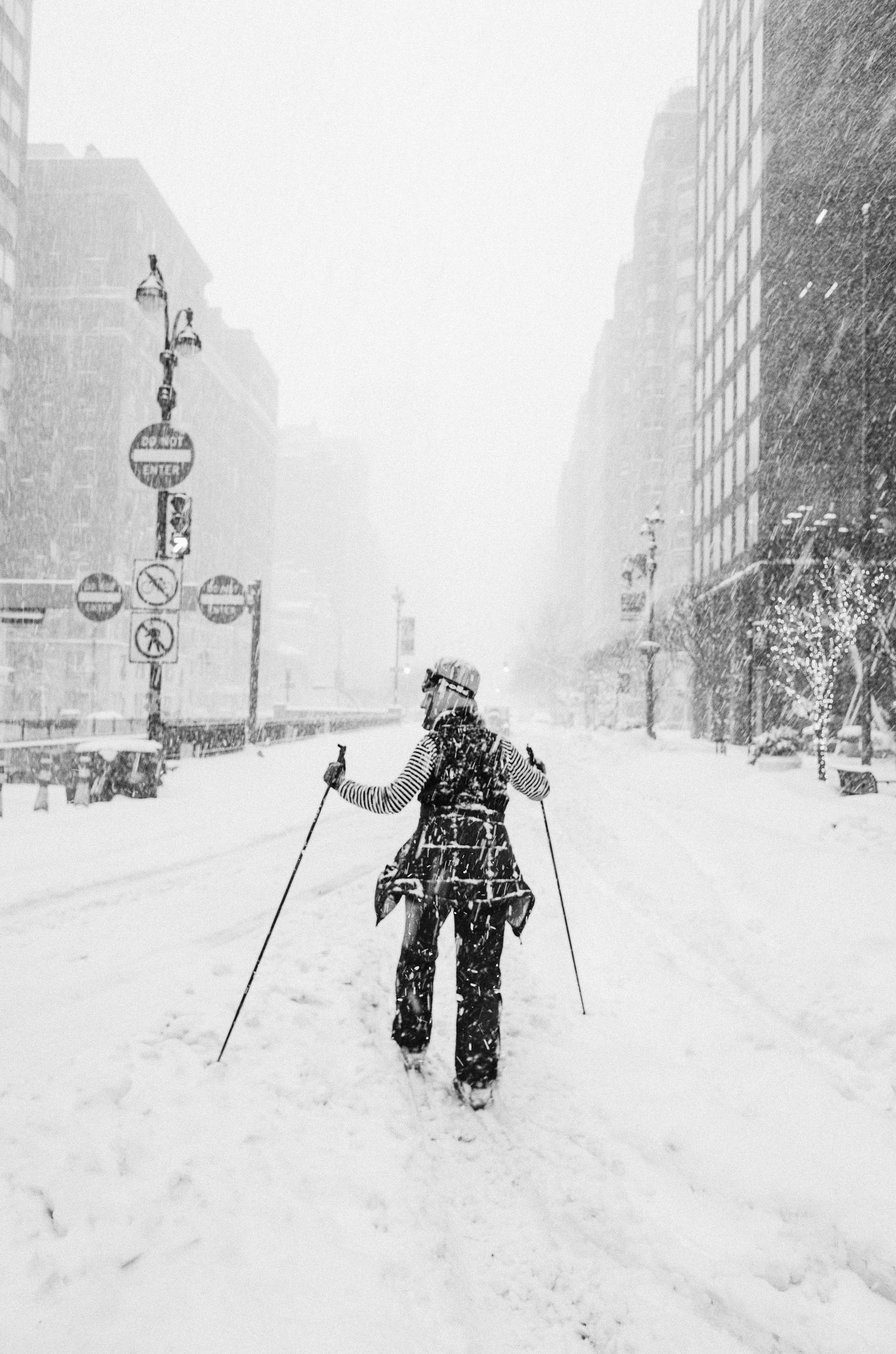 A person cross-country skiing down a snowy city street during a winter storm in an urban setting.