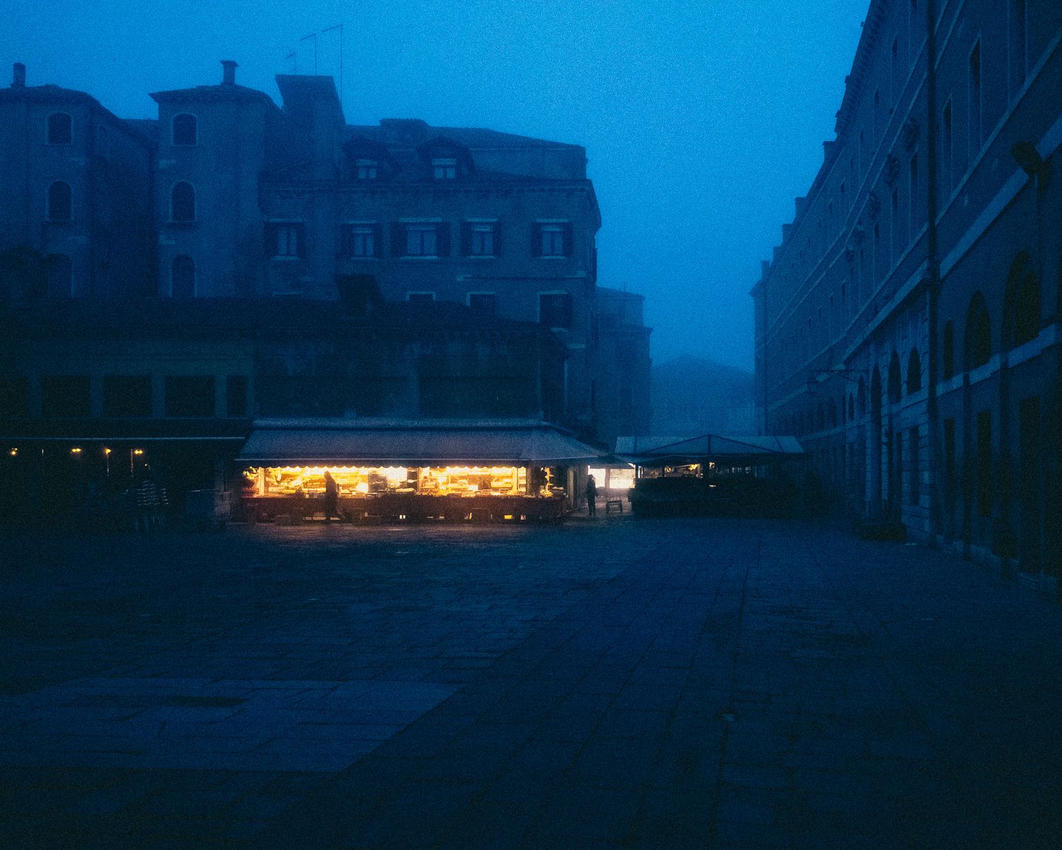 Panoramic view of city buildings at dusk with dark blue sky and distant lights creating a moody atmosphere.