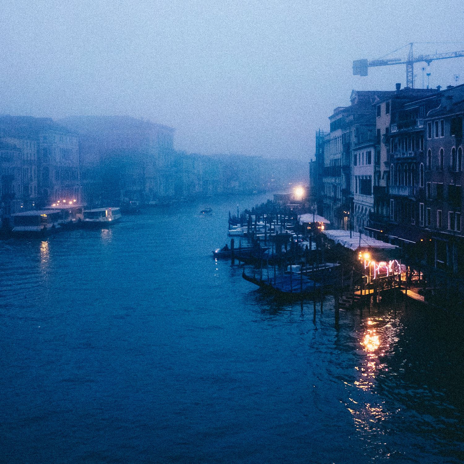 Foggy evening view of Venice's Grand Canal with illuminated buildings and boats reflecting on misty waters.