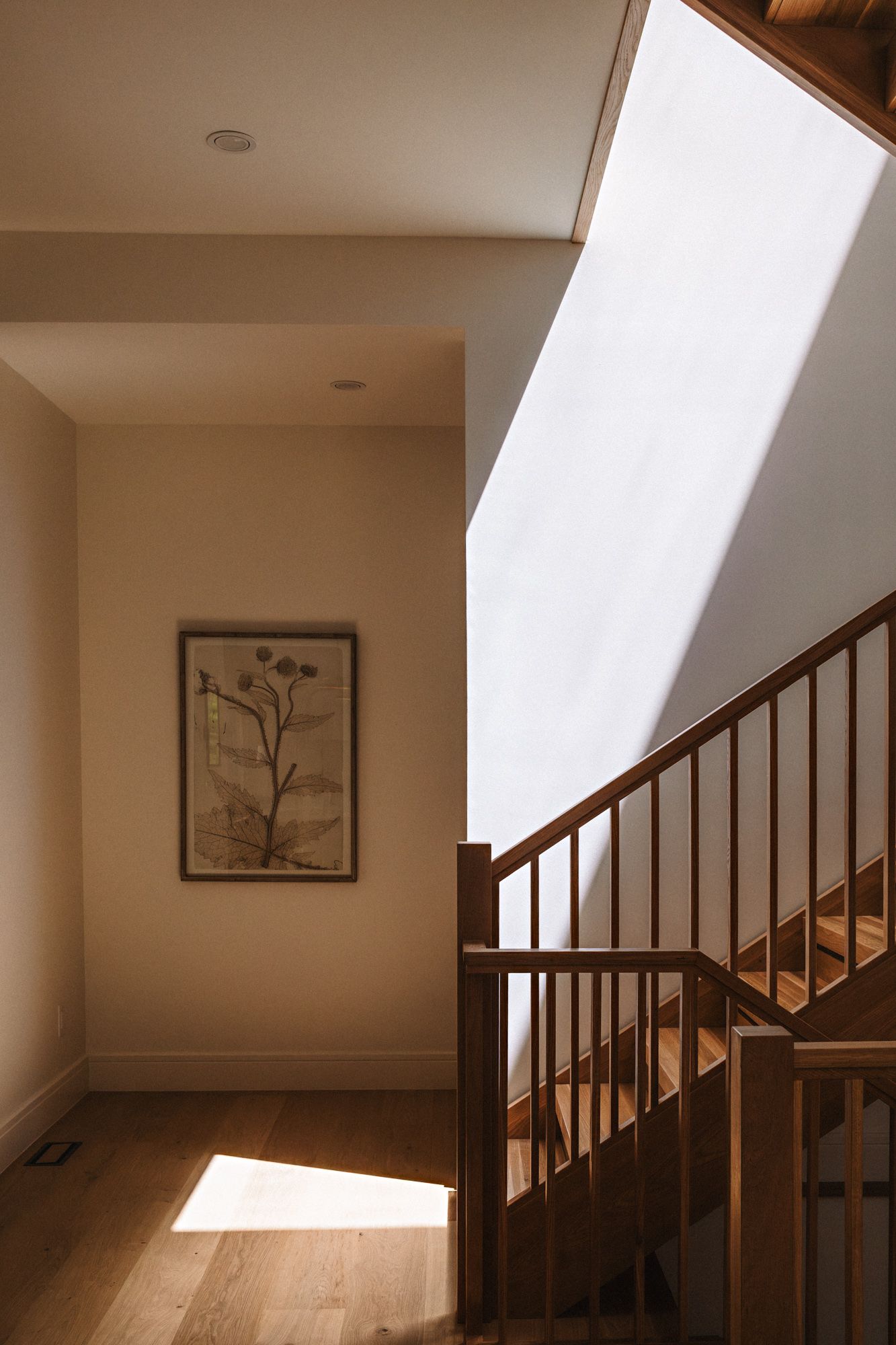 Sunlit hallway with wooden staircase, warm hardwood floors, and botanical framed art on cream walls.