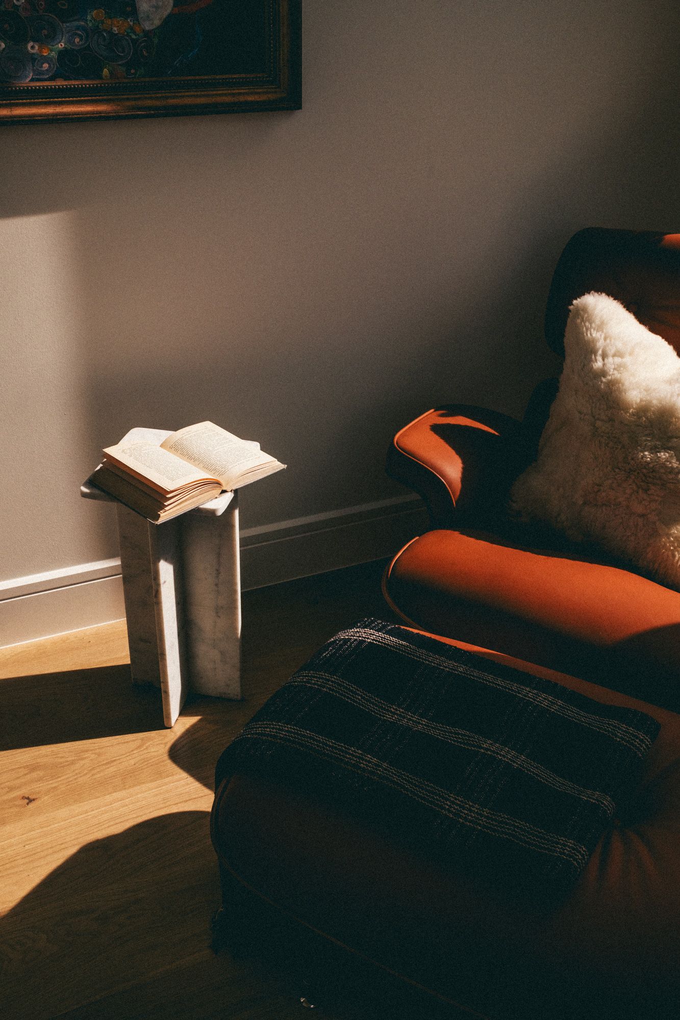 Cozy reading nook with an orange armchair, plaid ottoman, open book on a marble side table in warm sunlight.