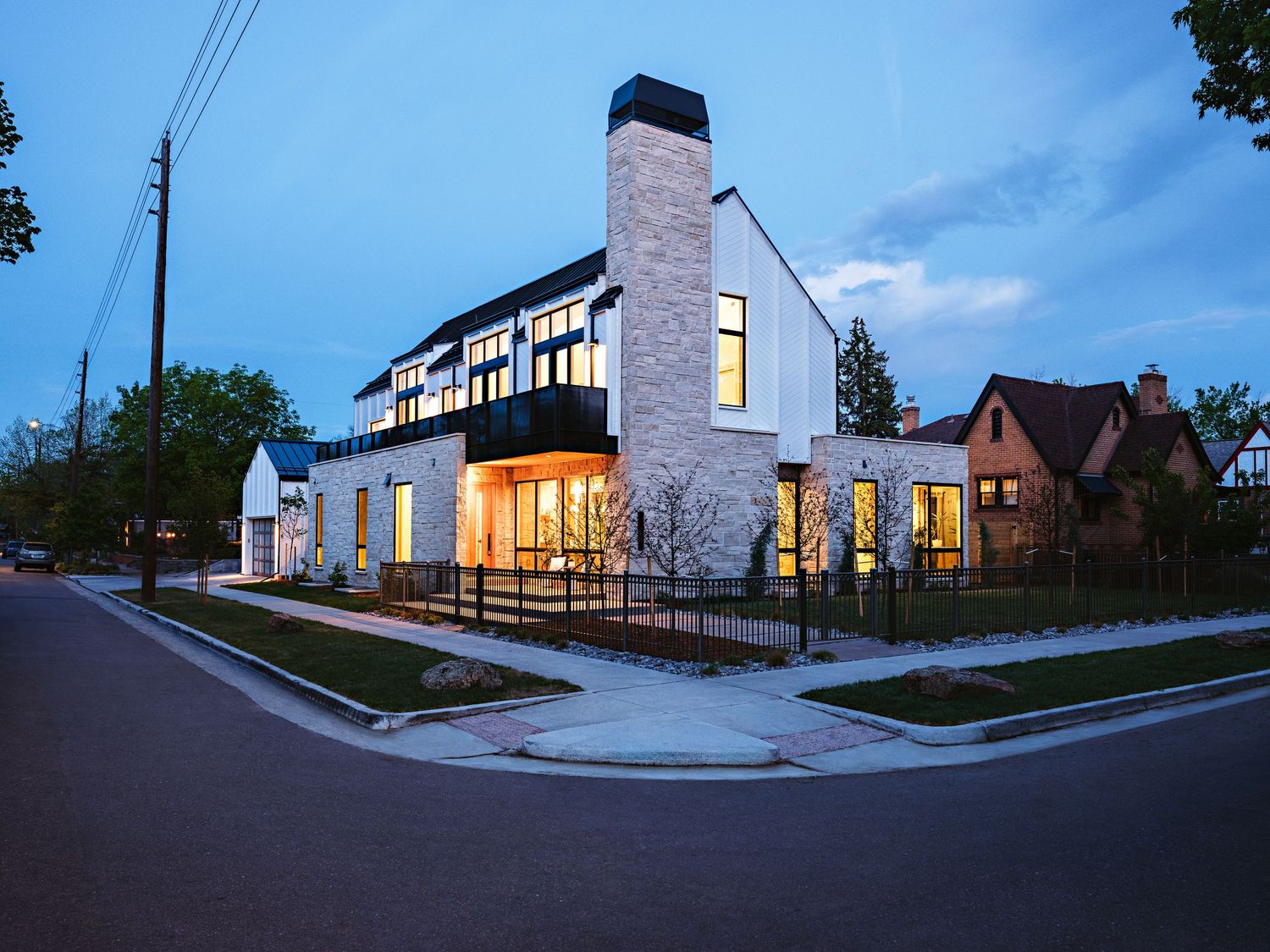 Modern two-story white stone home with large windows and chimney photographed at dusk on a residential street corner.