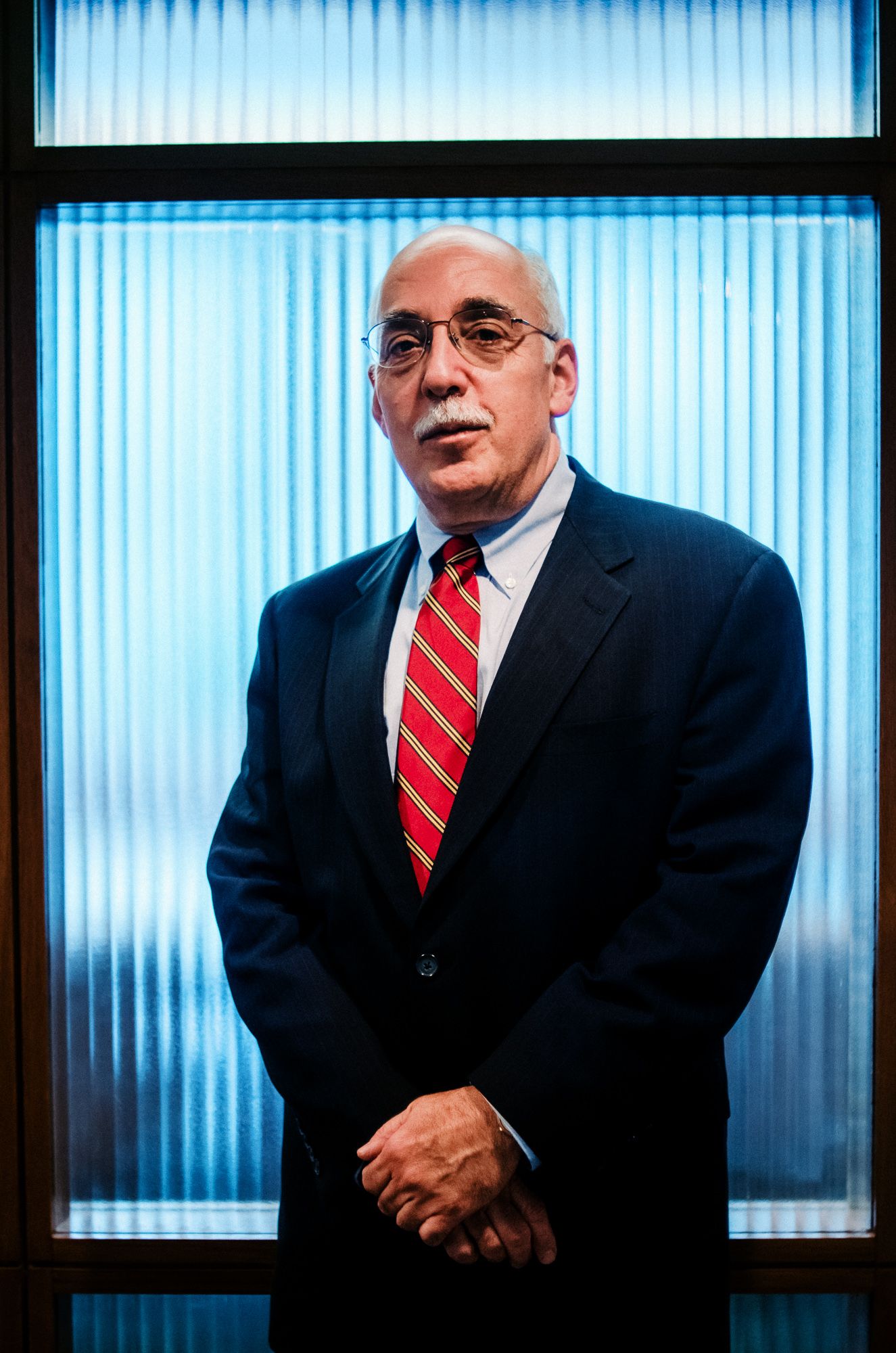 Professional portrait against blue striped background wearing dark suit and red tie.