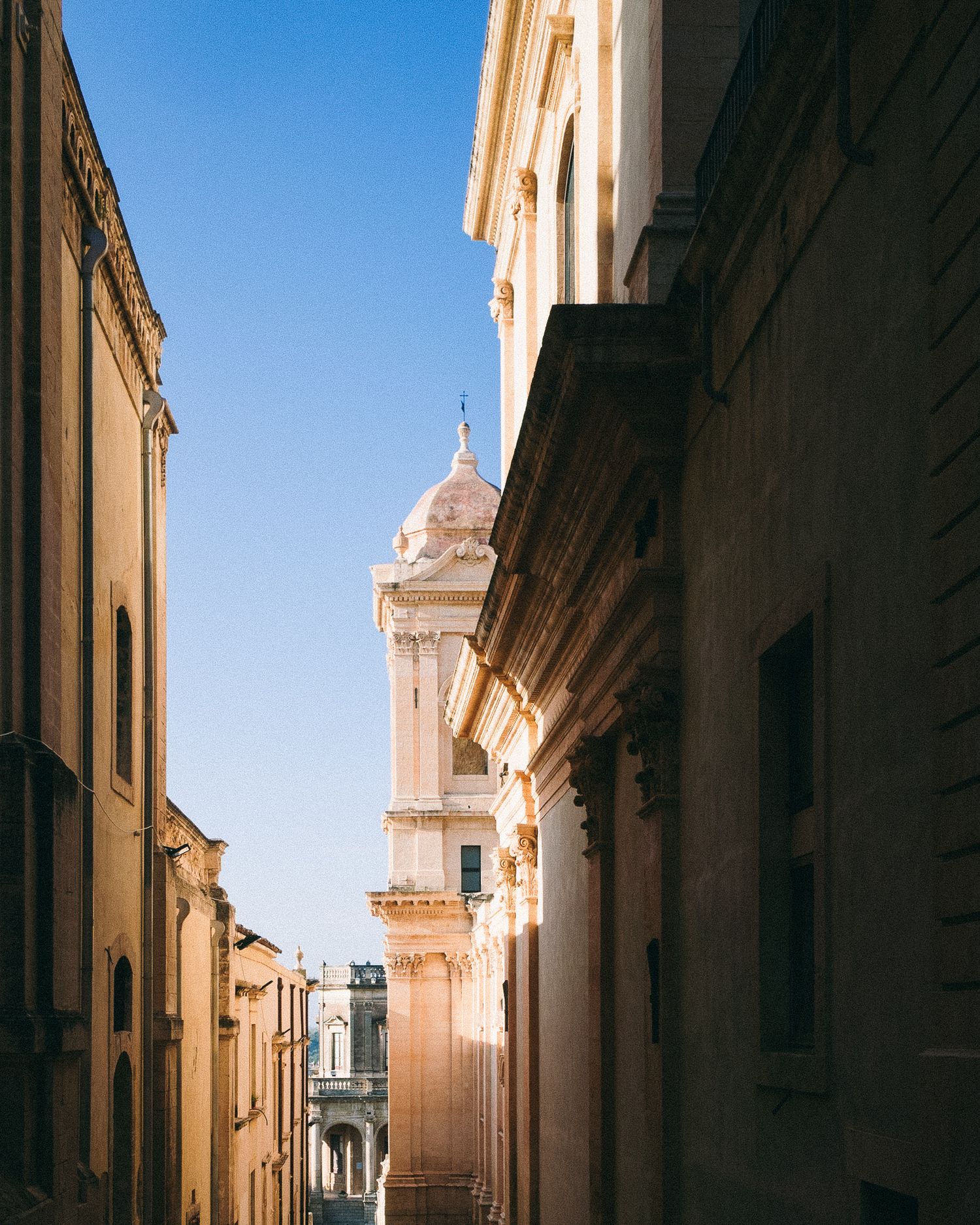 Narrow alley in Mediterranean town with historic church tower and beige stone buildings under bright blue sky.