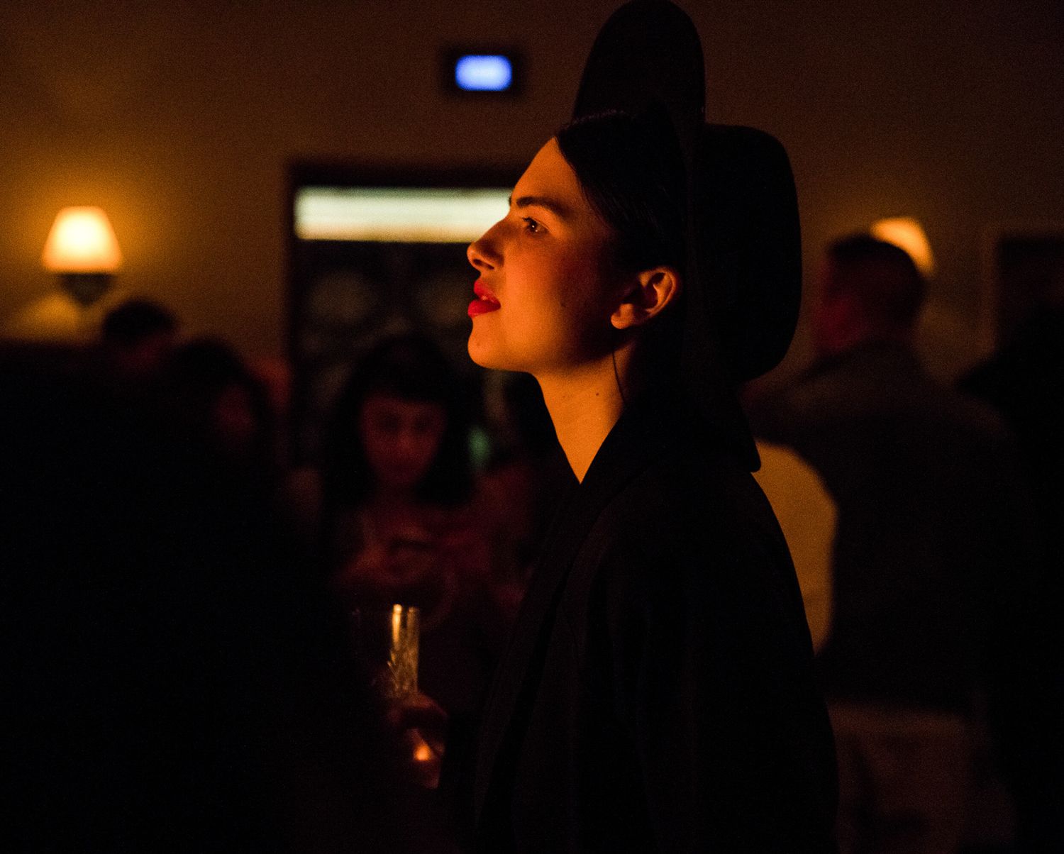 Woman in black outfit and hat holding a drink at a dimly lit evening social gathering.