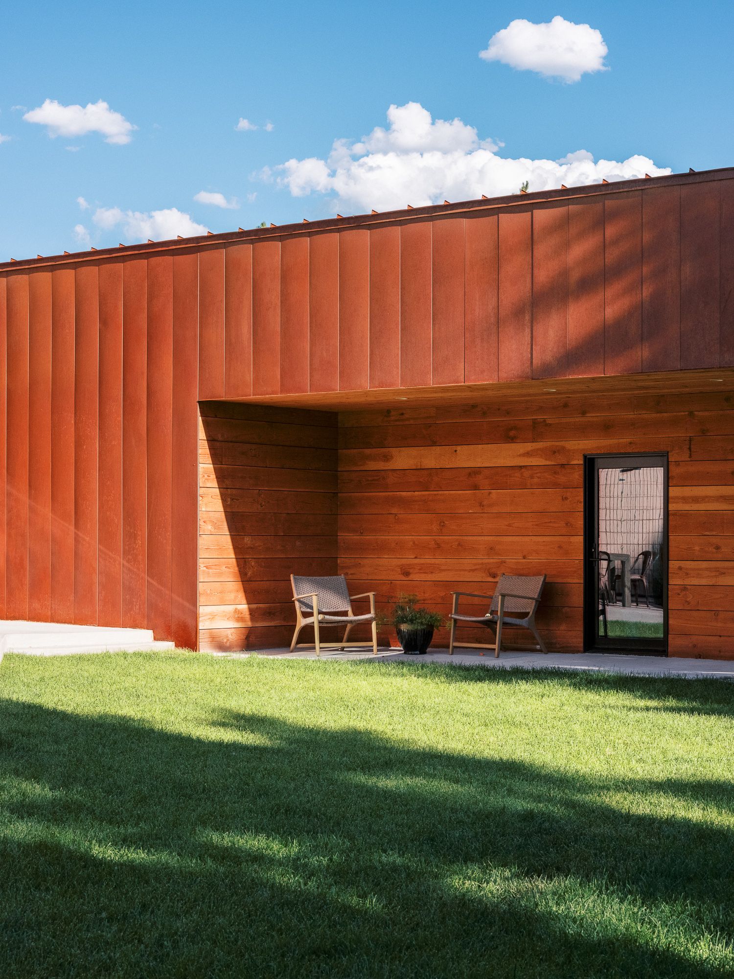 Long rust-colored metal wall with geometric patterns stretching across a green lawn under blue sky.
