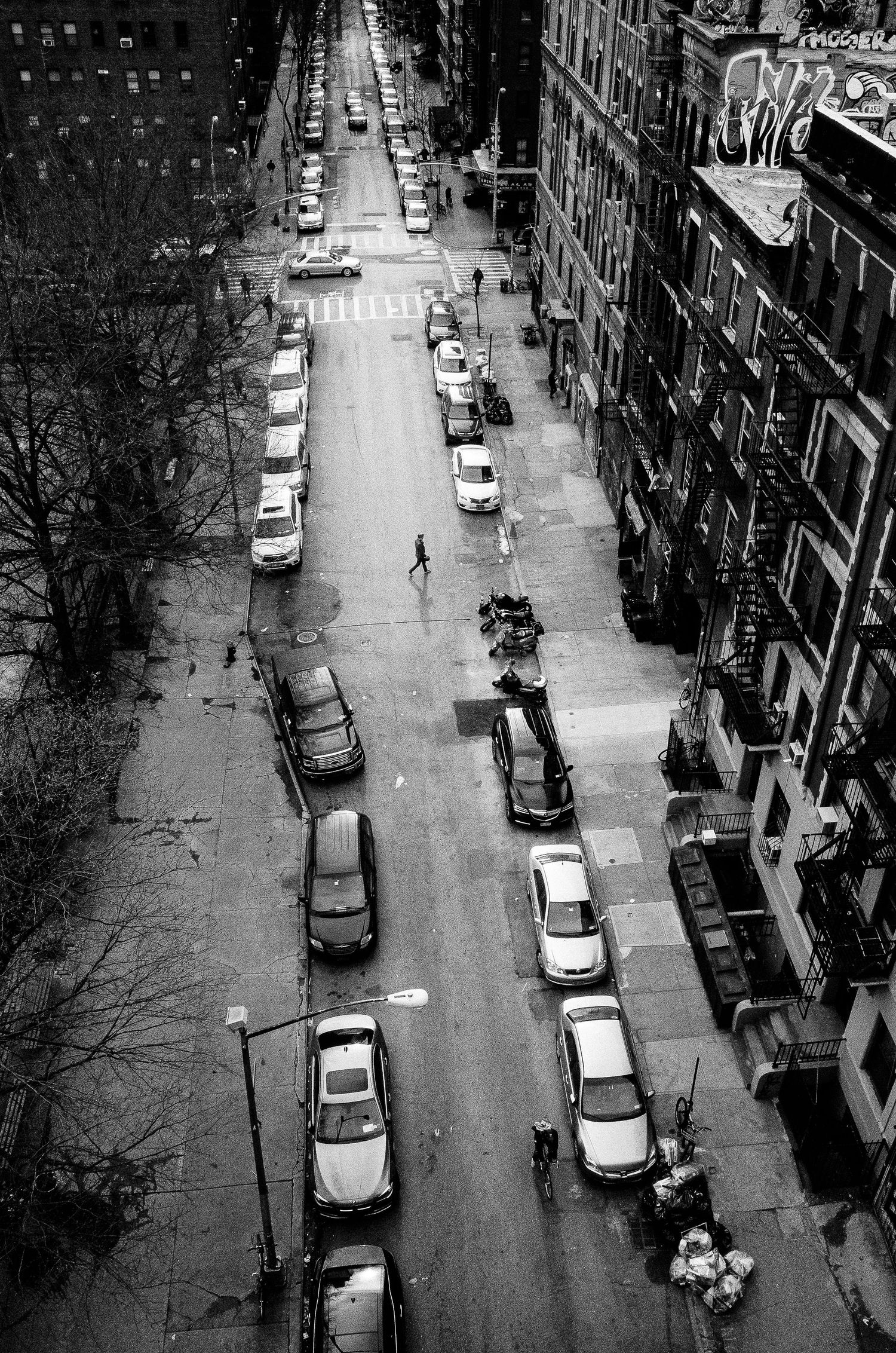 Black and white aerial view of a city street with parked cars and pedestrians walking along apartment buildings.