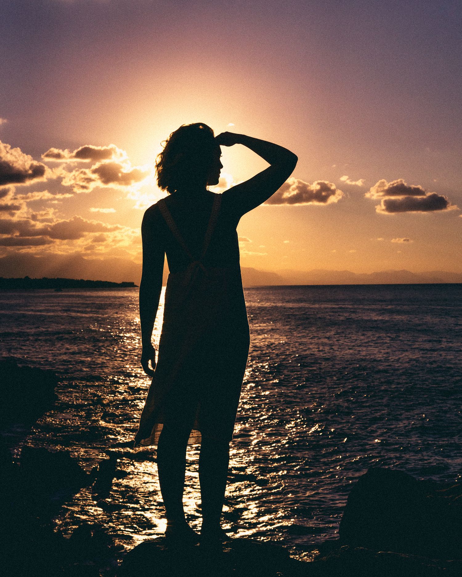 Silhouetted figure stands at ocean's edge during golden sunset creating dramatic shadow against sparkling water.