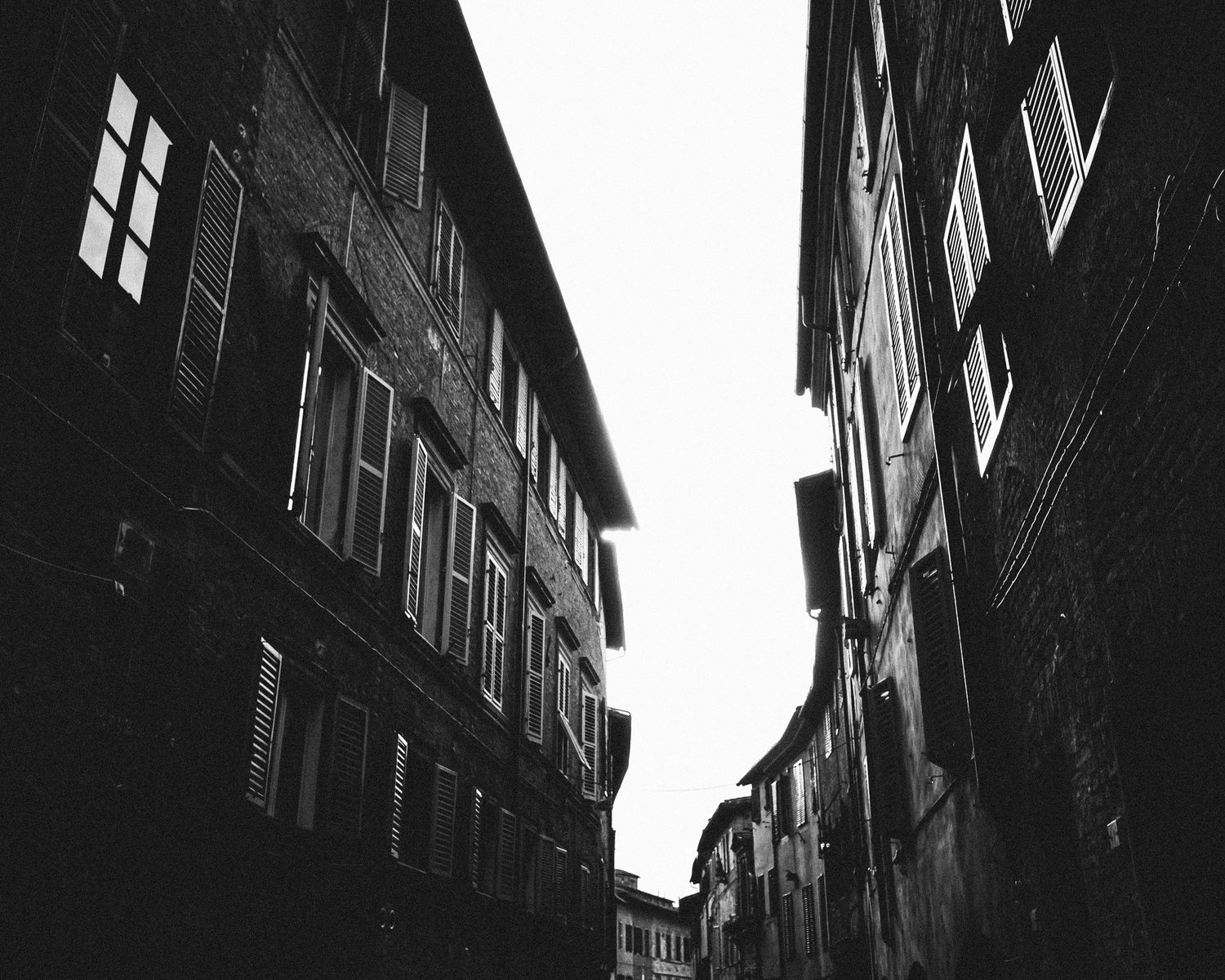 A dramatic black and white view of a narrow European alleyway with tall buildings and illuminated windows.