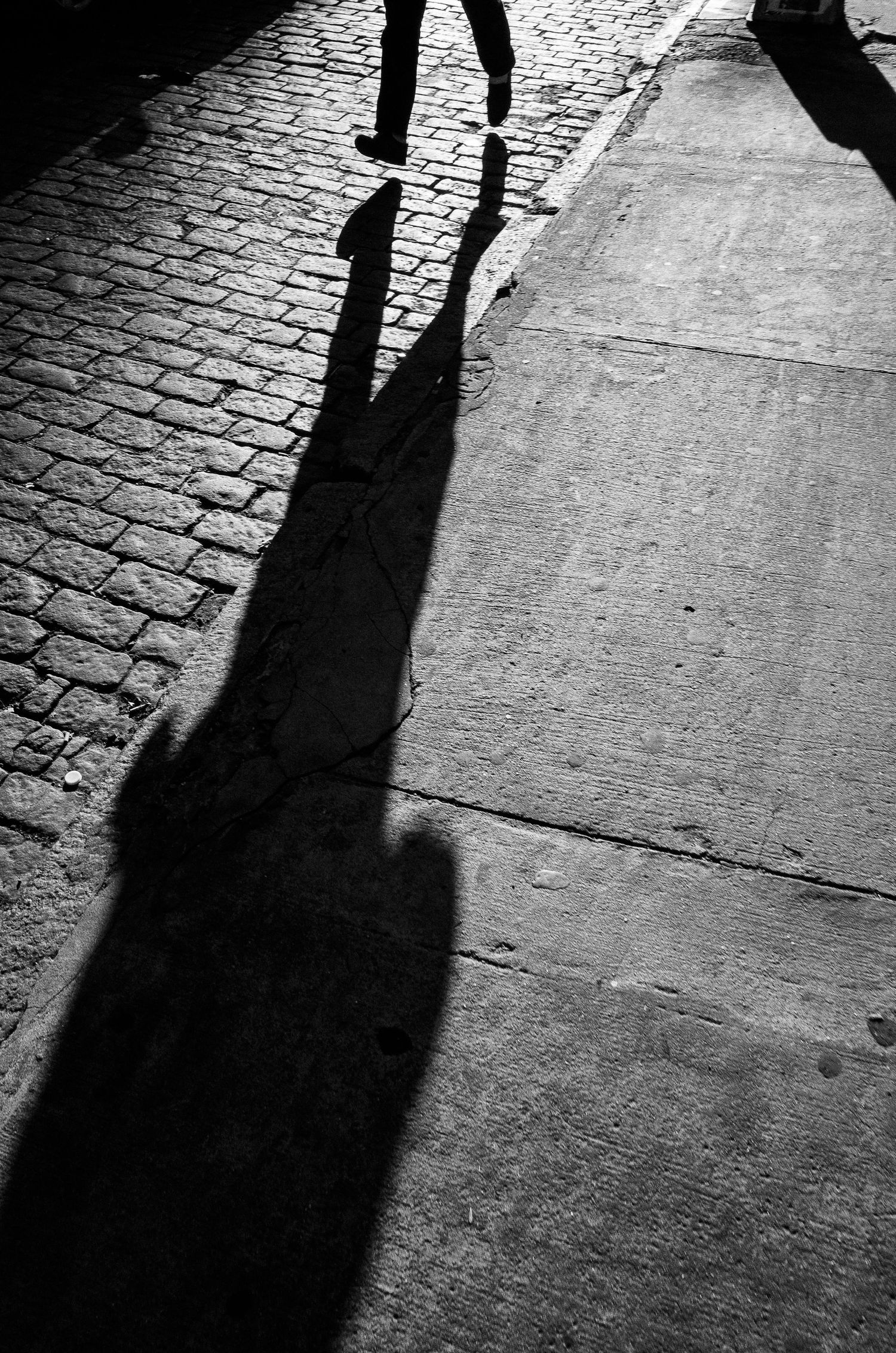 A sequence of black shadows cast on cobblestone pavement shows a person walking across the frame.