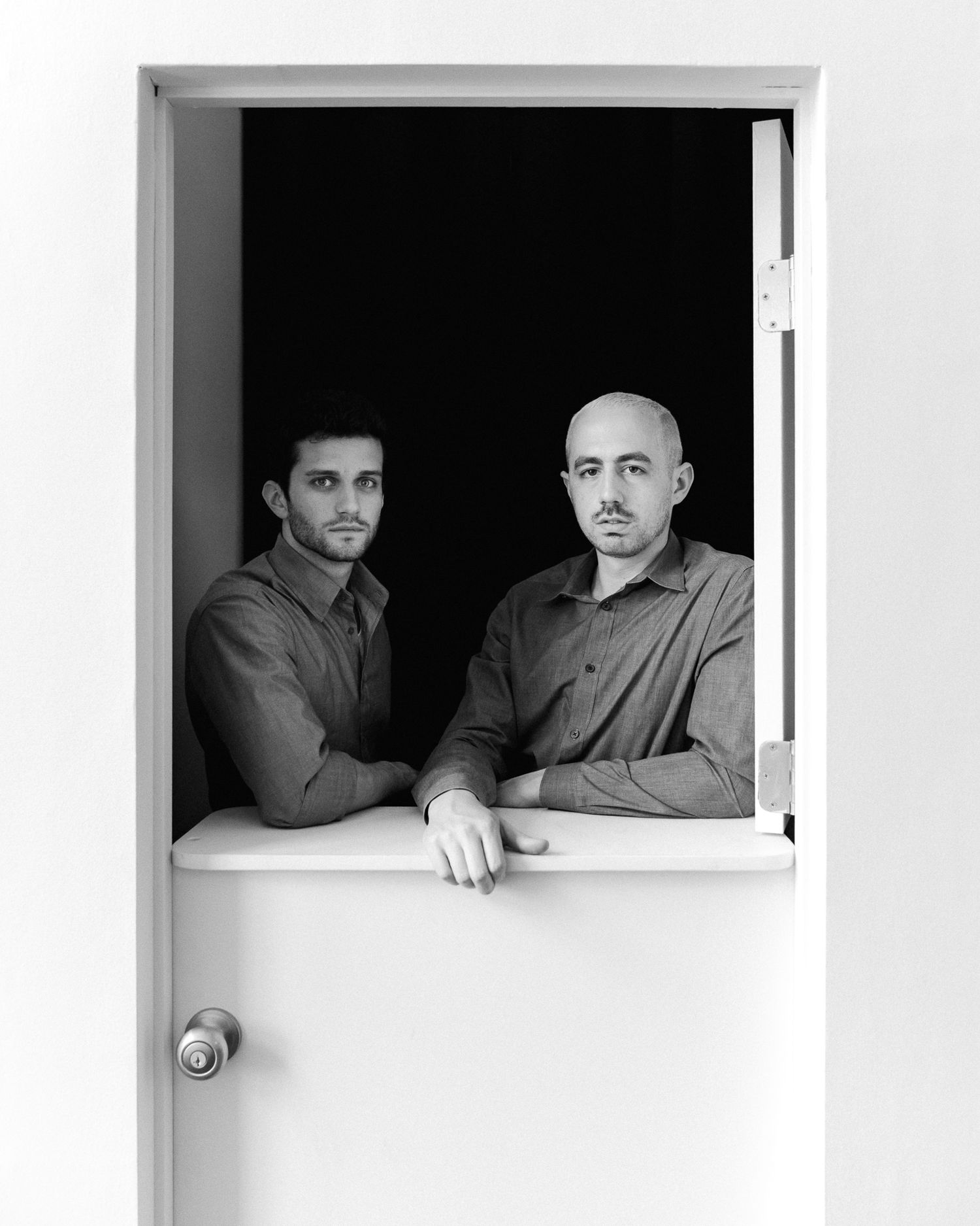 Two figures framed in a white doorway against a dark background in black and white portrait.