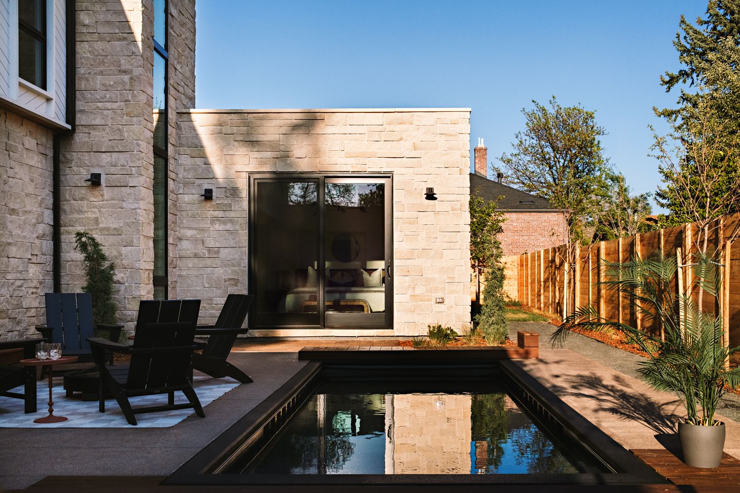 Modern home exterior with stone facade, black pool, wooden deck, and patio chairs in a landscaped backyard.