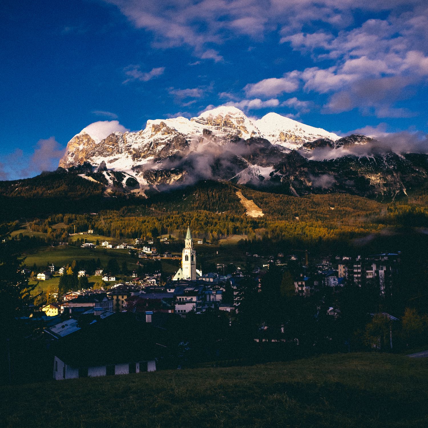 Panoramic view of a mountain village nestled beneath snow-capped peaks with dramatic clouds at sunset in the Dolomites.
