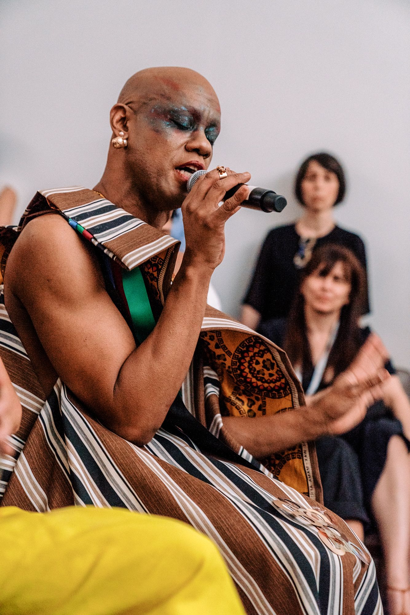Bald performer with dramatic eye makeup singing into microphone wearing striped traditional garment at live cultural event.