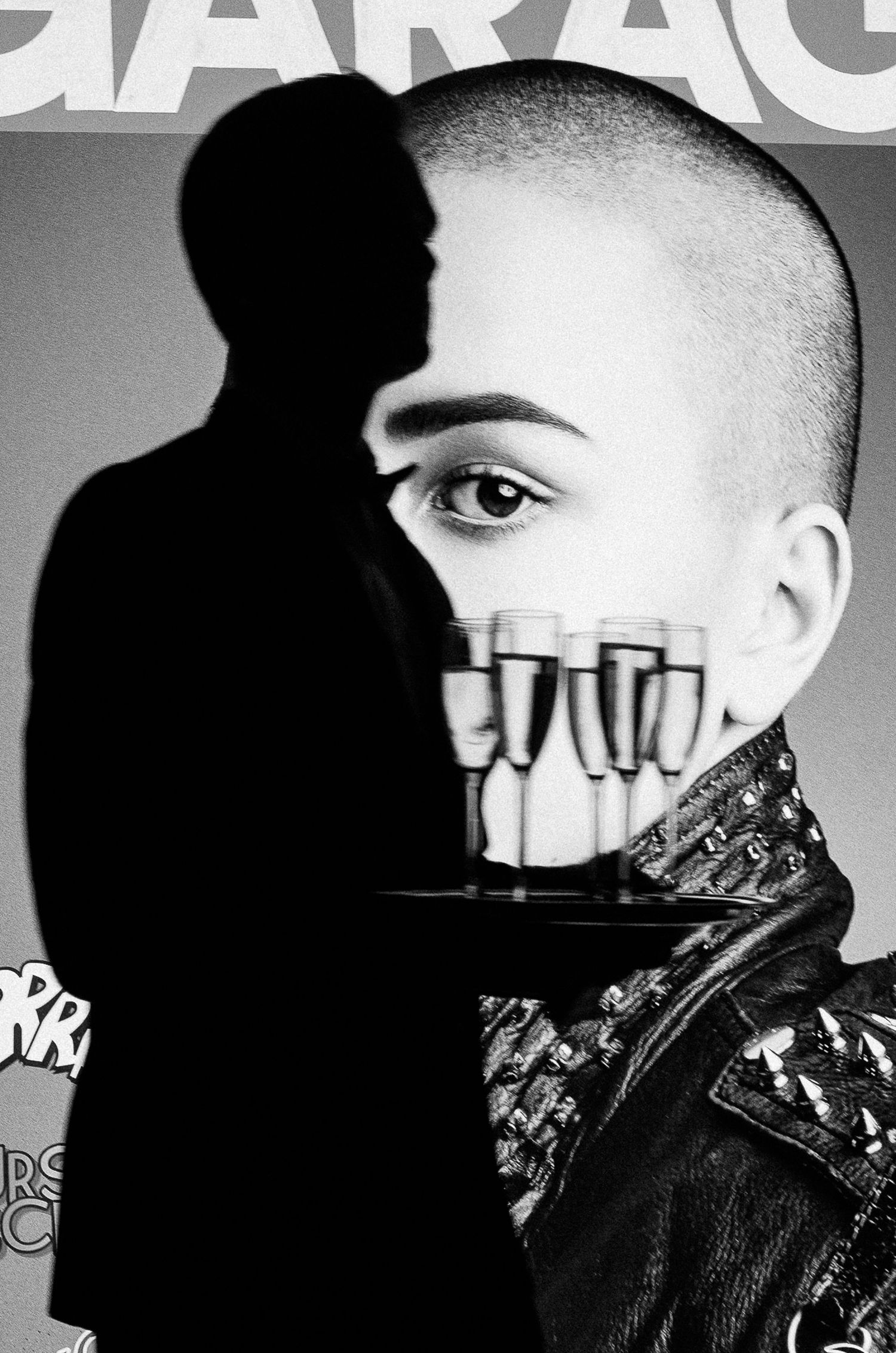 Black and white artistic portrait with face partially covered by champagne glasses, wearing leather jacket and shaved head.