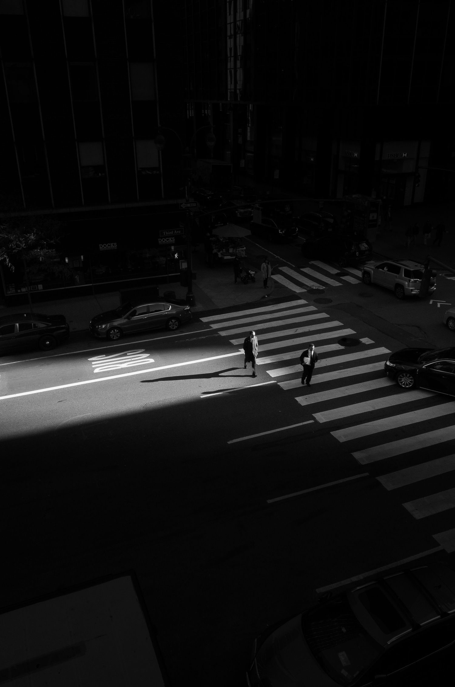 Two silhouettes cross an illuminated crosswalk at night in a dark urban street scene.