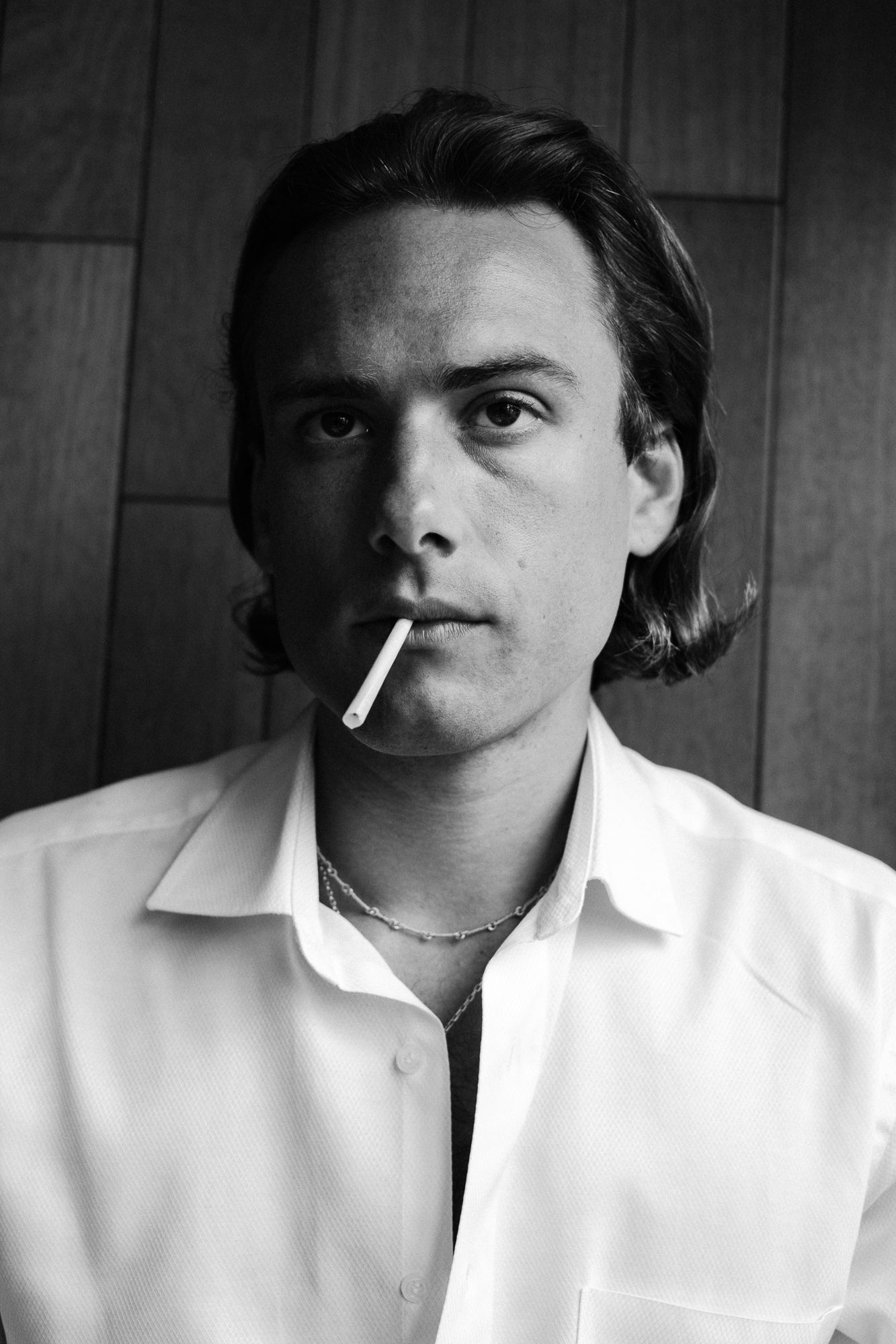 Black and white portrait with cigarette wearing white shirt against wooden paneled wall.