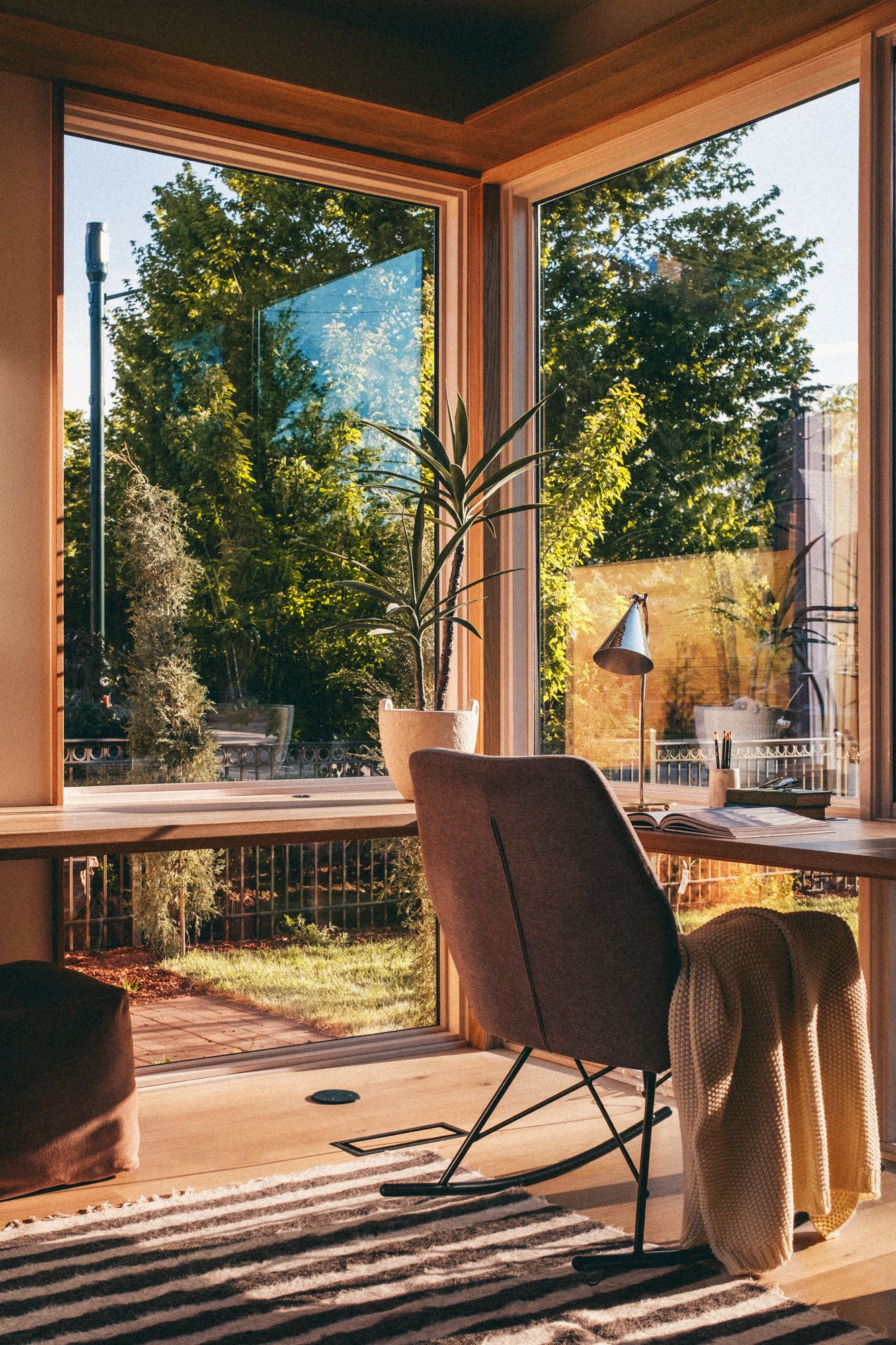 Sunlit home office corner with floor-to-ceiling windows overlooking lush garden, brown chair, and potted plant on wood desk.